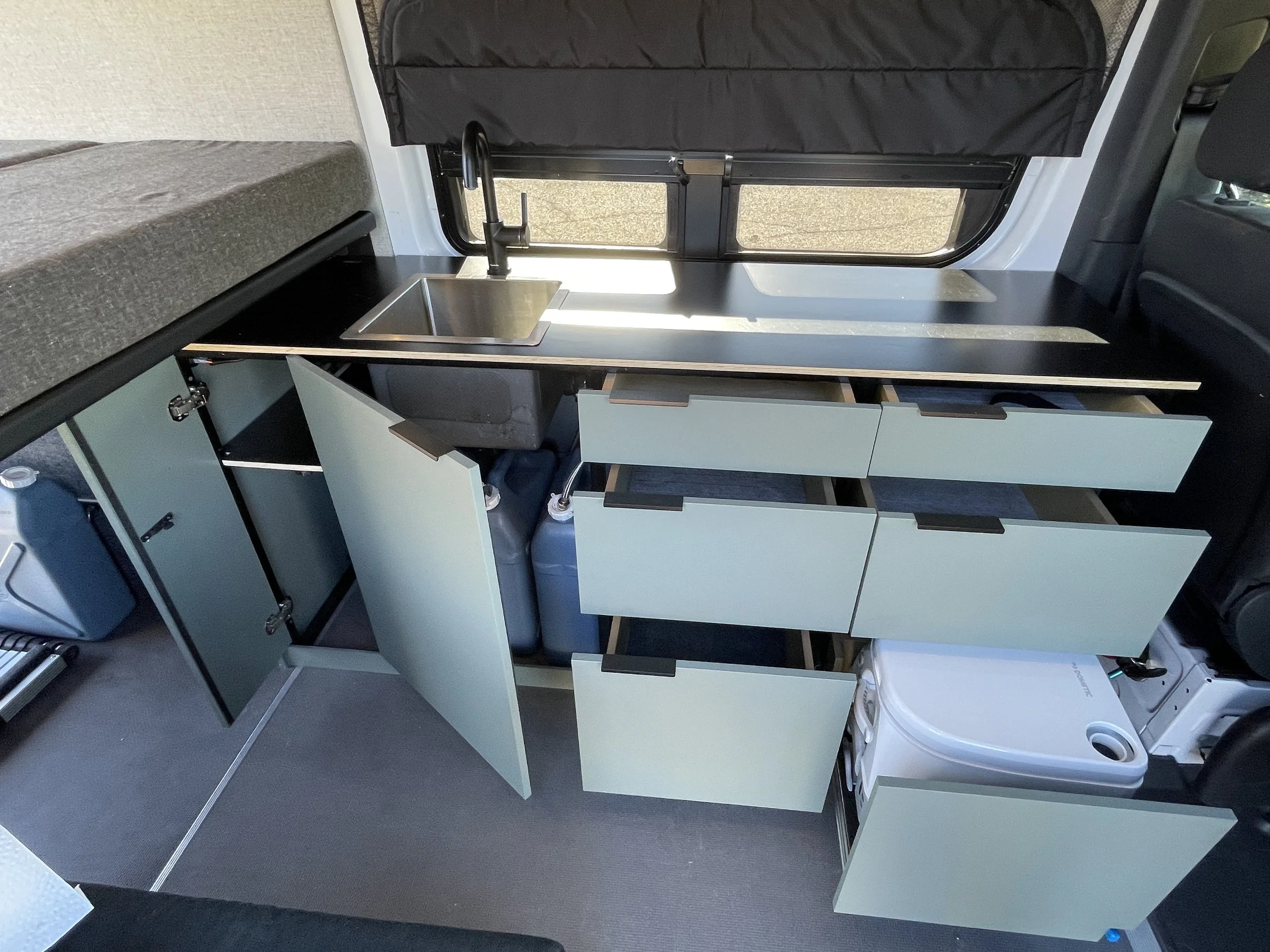 Interior of a camper van showing a kitchenette with open cabinets, drawers, a small sink, and a portable toilet.