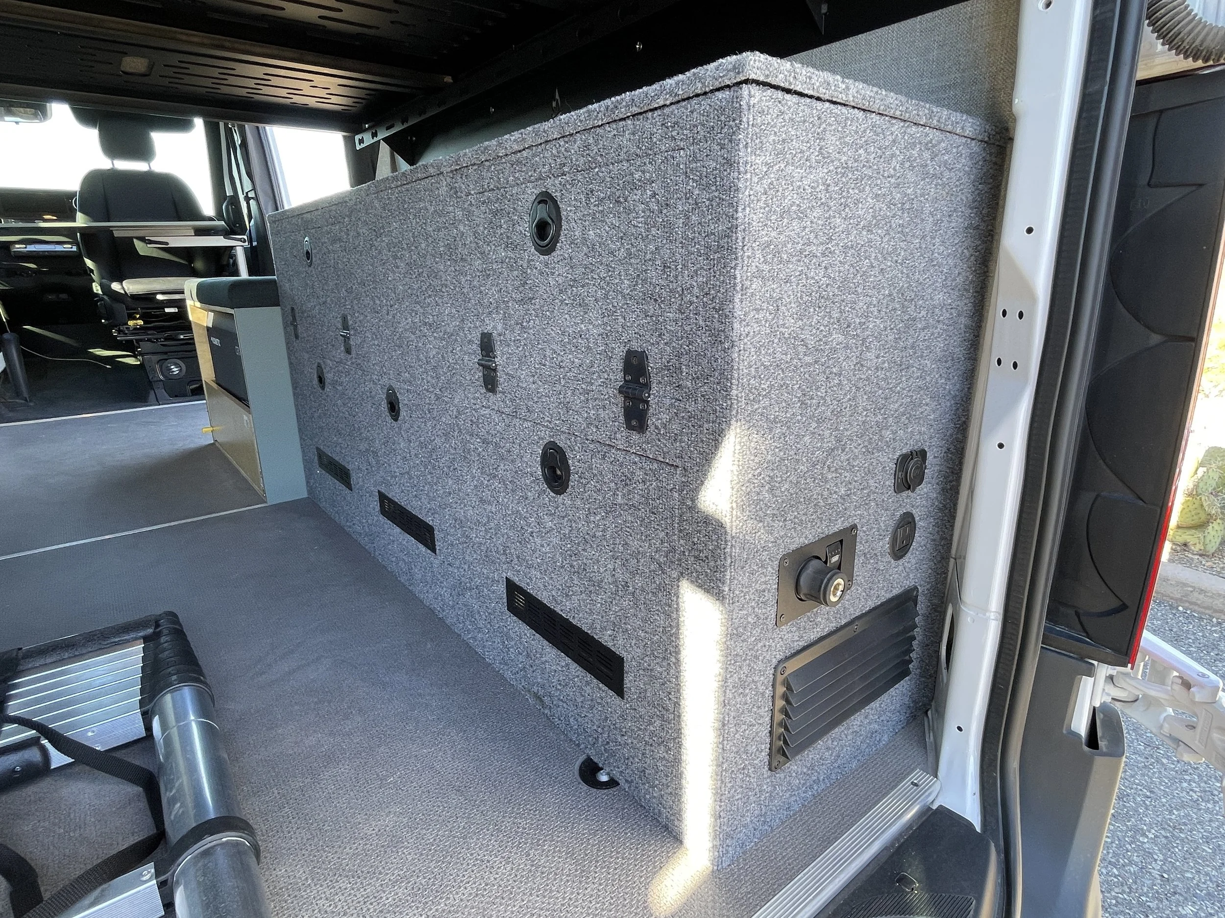Interior of a custom van conversion showing a built-in gray carpeted storage cabinet, various circular vents, and utility outlets. A collapsible ladder is resting on the floor.