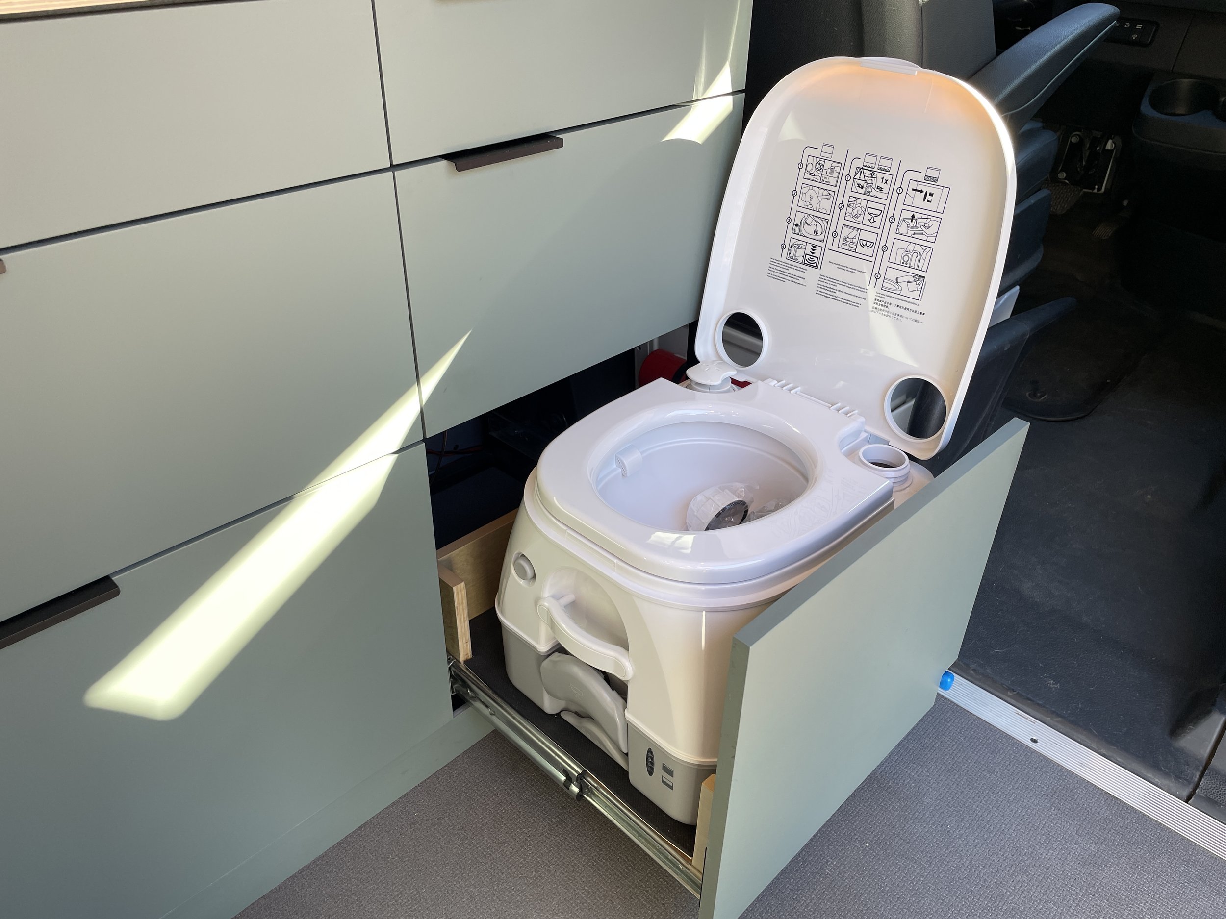 Portable toilet installed in a compartment of a van conversion, with a pull-out drawer mechanism, and instructions on the lid.
