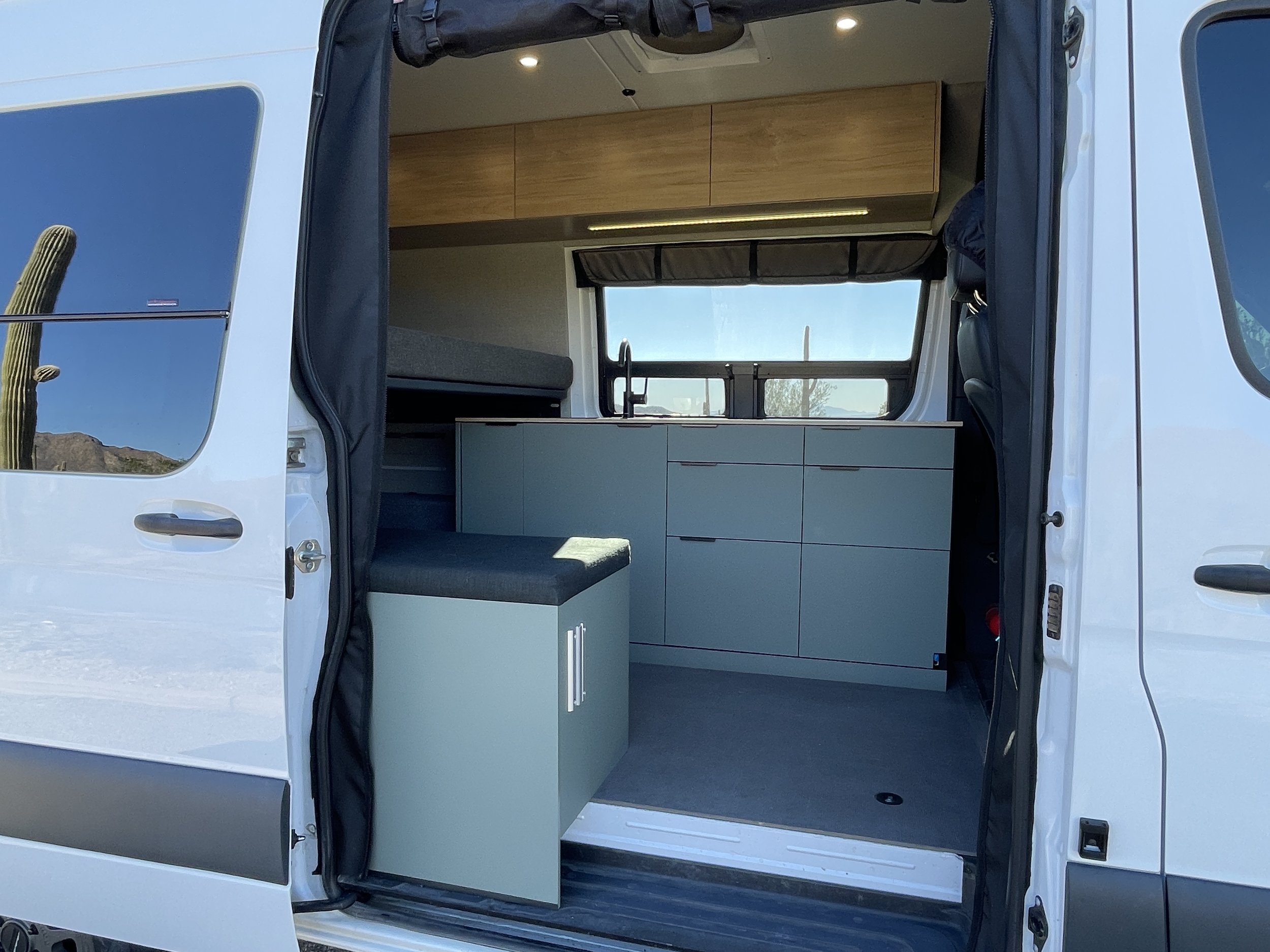Interior view of a camper van with open sliding door, featuring a kitchenette with cabinets, a countertop, and seating. The surrounding landscape includes a cactus visible through the window.
