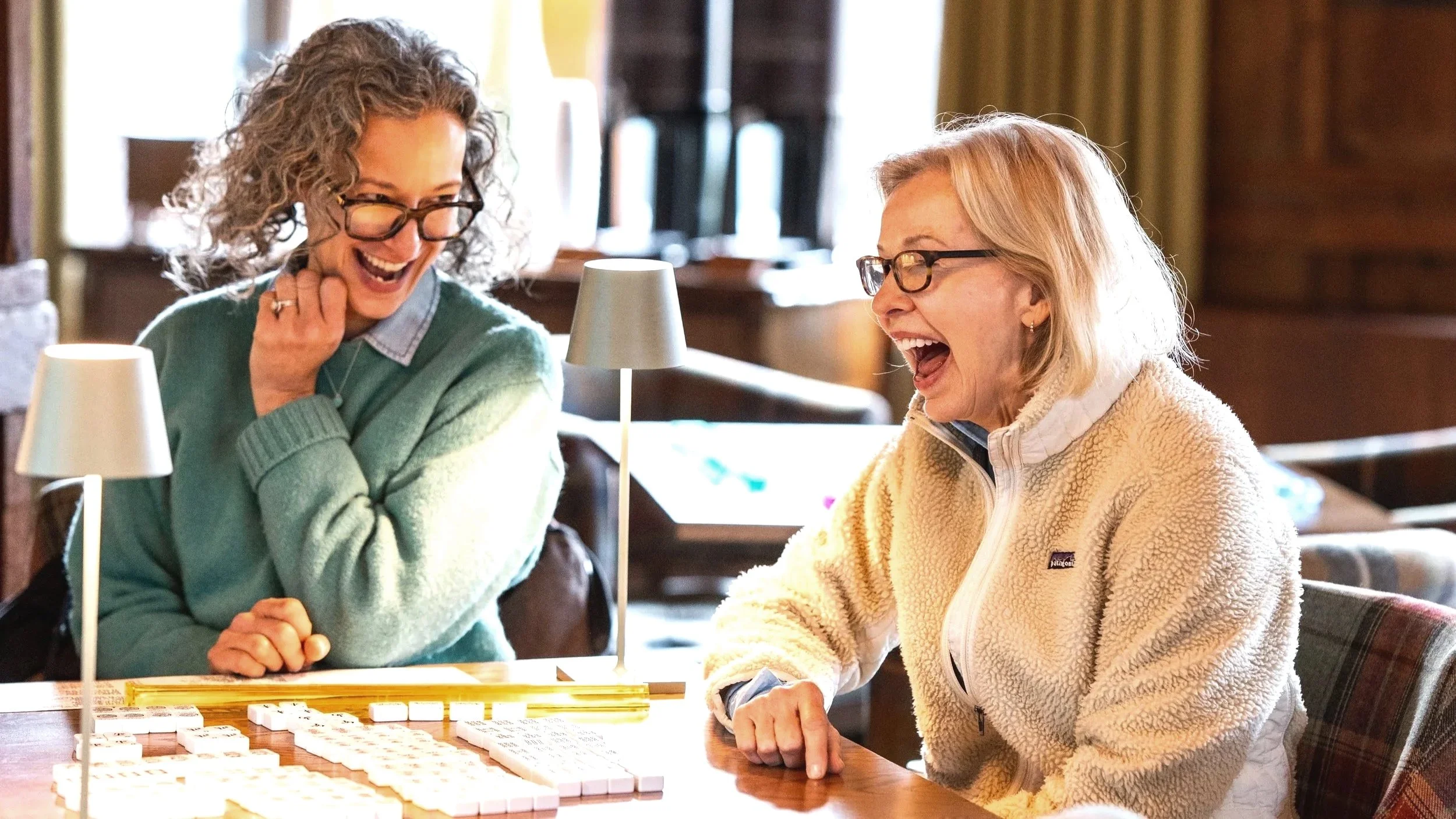 Two women laughing and playing a game of Mah Jongg at a wooden table with table lamps in a cozy, warmly lit room.
