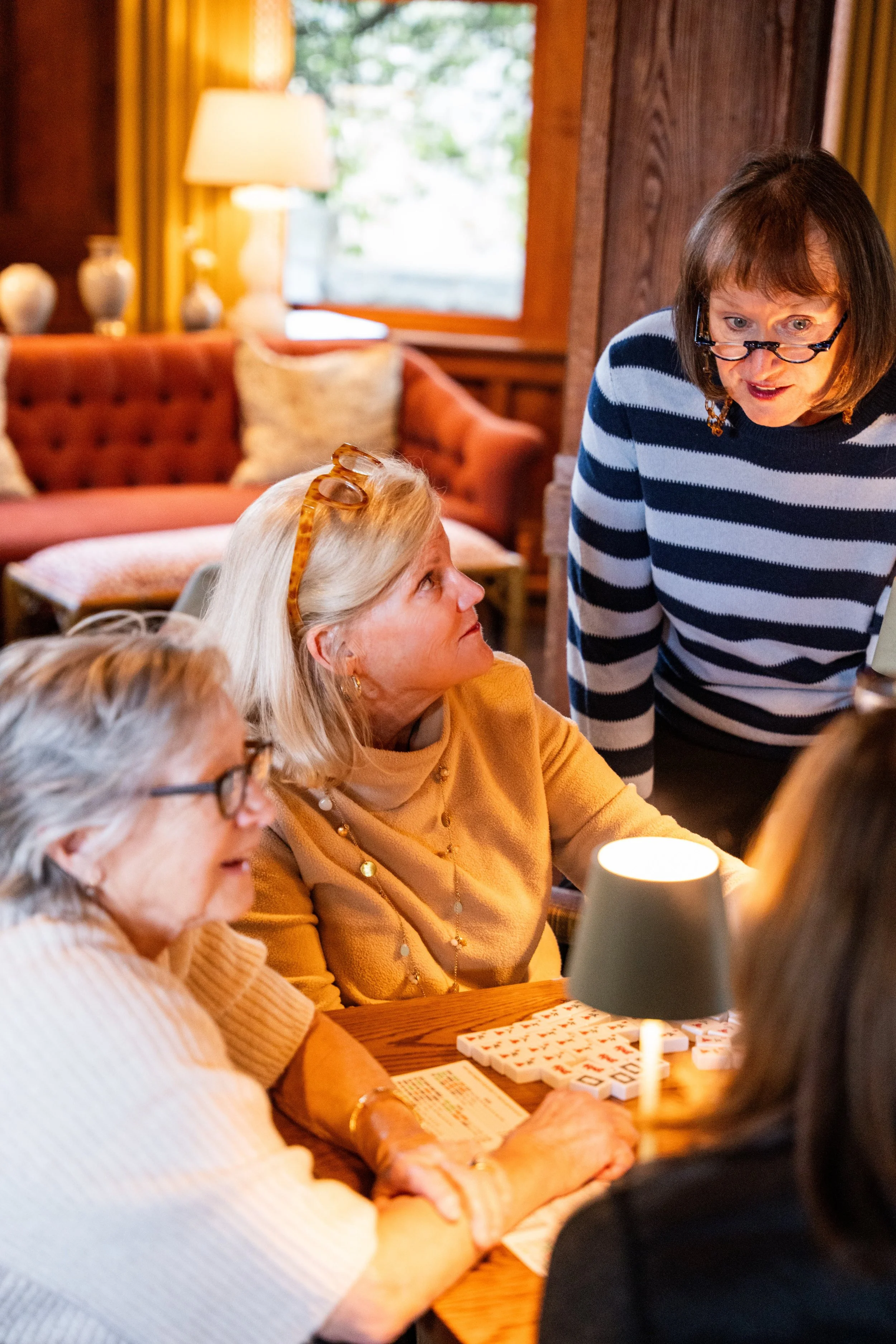 A group of women playing a game of Mah Jongg in a cozy, warmly lit living room.