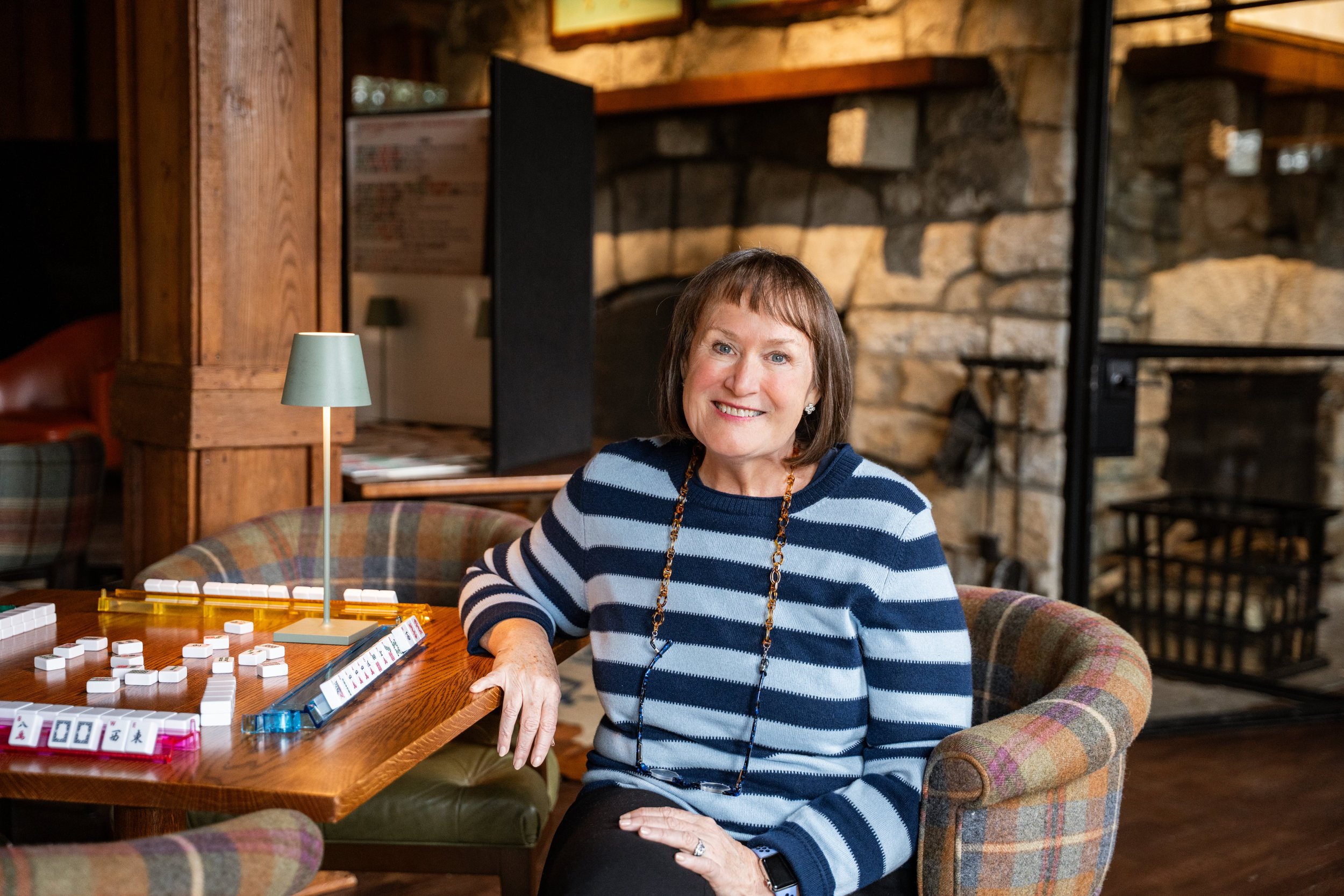 Mah Jongg teacher Dana Lange sits at a game table inside a cozy room with wooden walls and a stone fireplace.