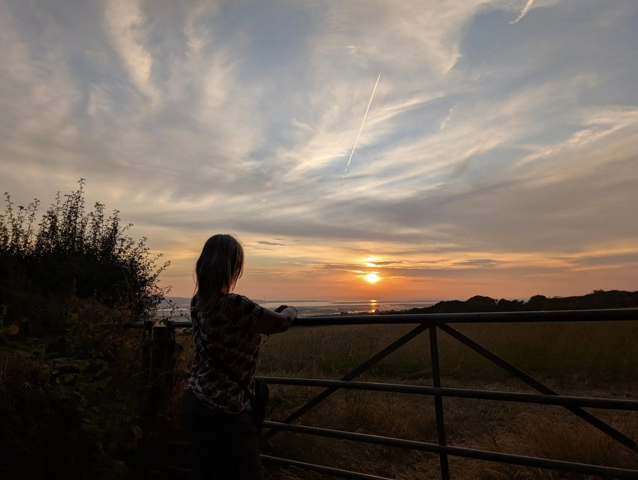 A young girl standing at a fence, watching the sunset over the ocean with clouds in the sky.