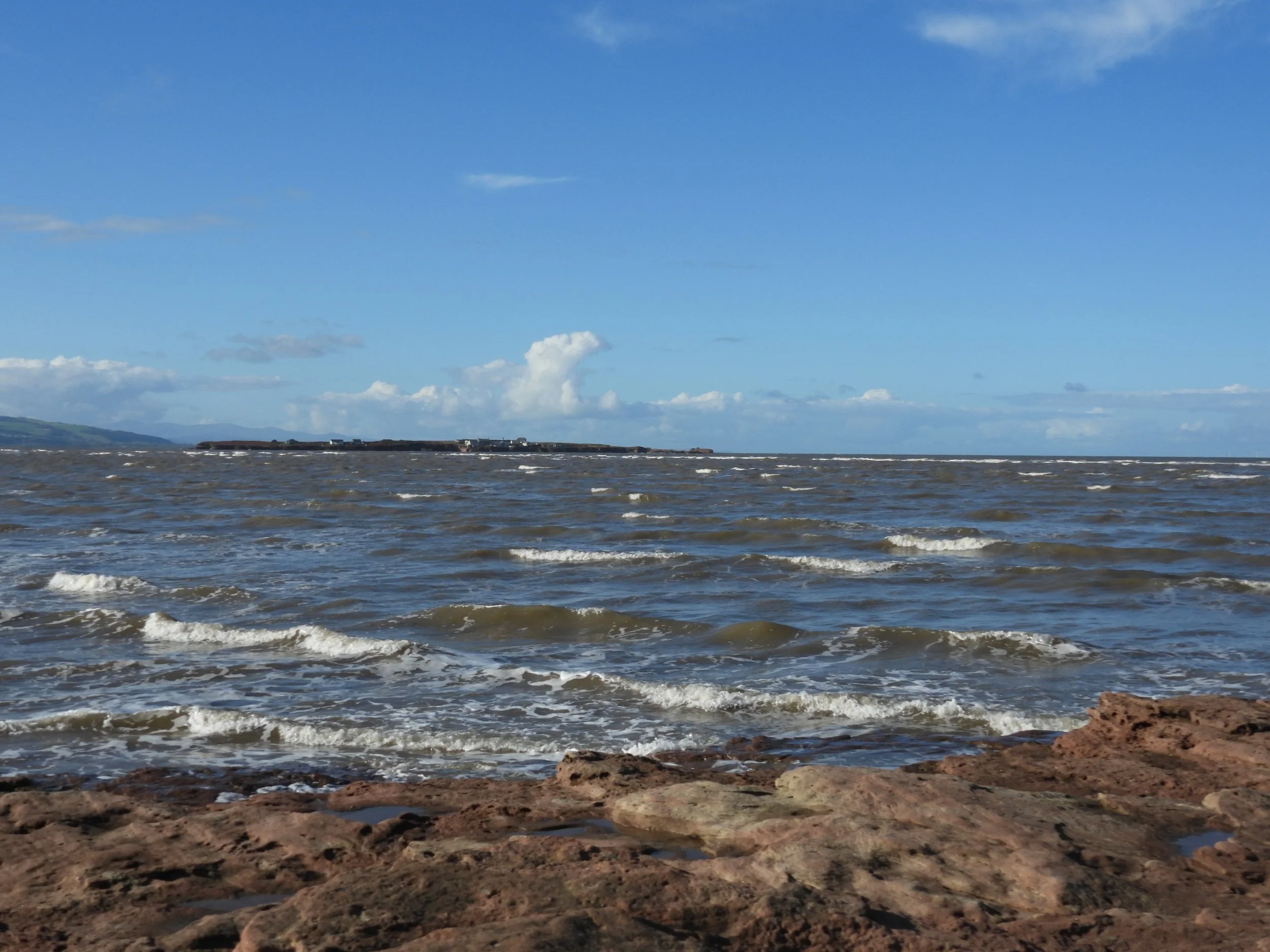 View of the ocean with waves lapping against the rocky shore, a distant landmass on the horizon, and partly cloudy blue sky overhead.