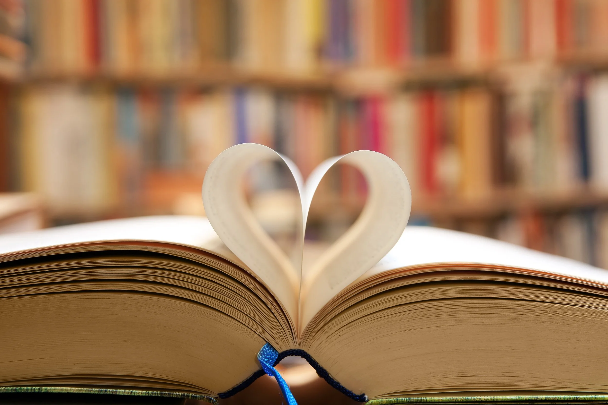 Open book with two pages folded into heart shape, against blurred bookshelf background.