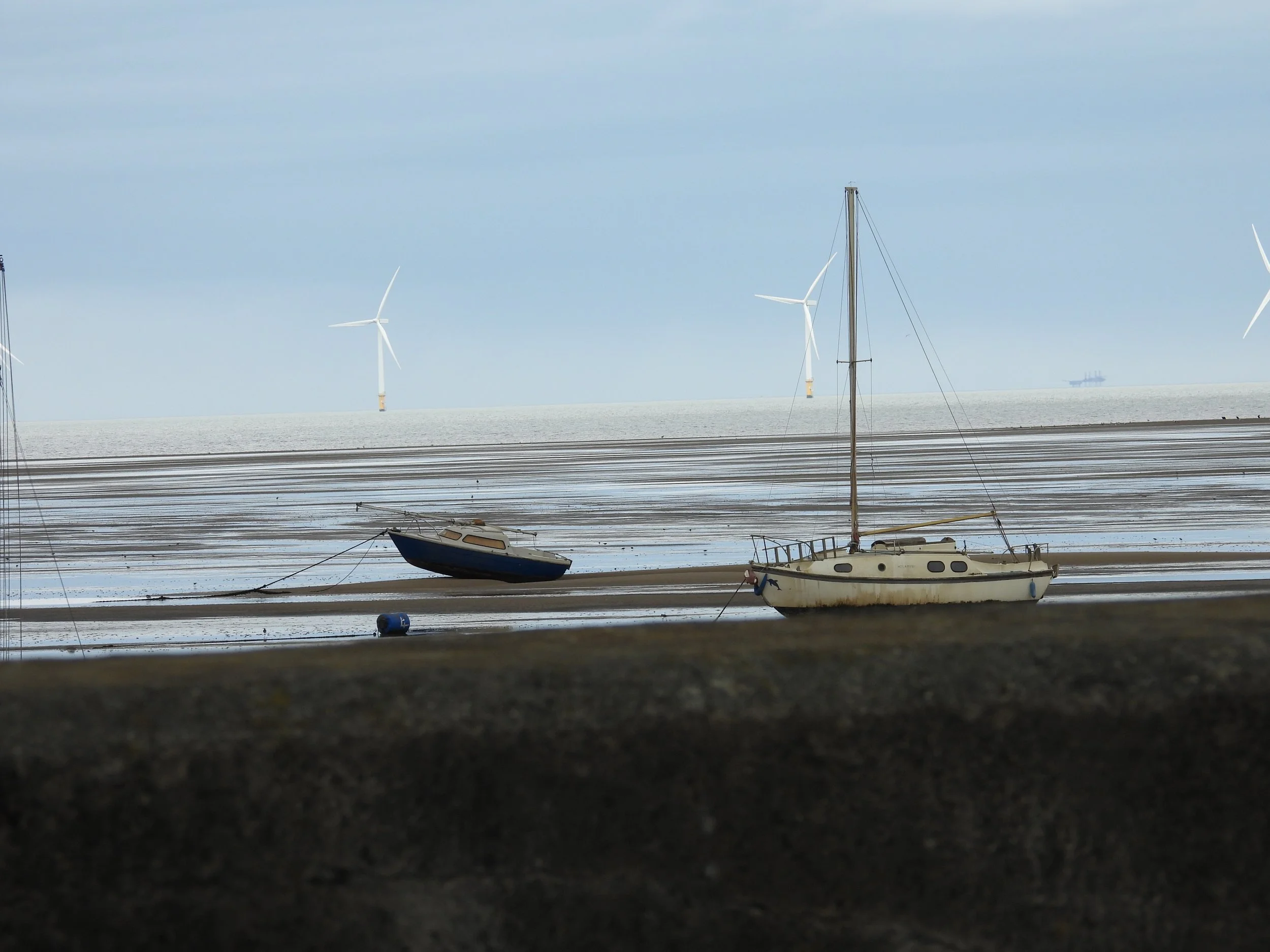 Two sailboats on a muddy shore with wind turbines in the background and an ocean in the distance.