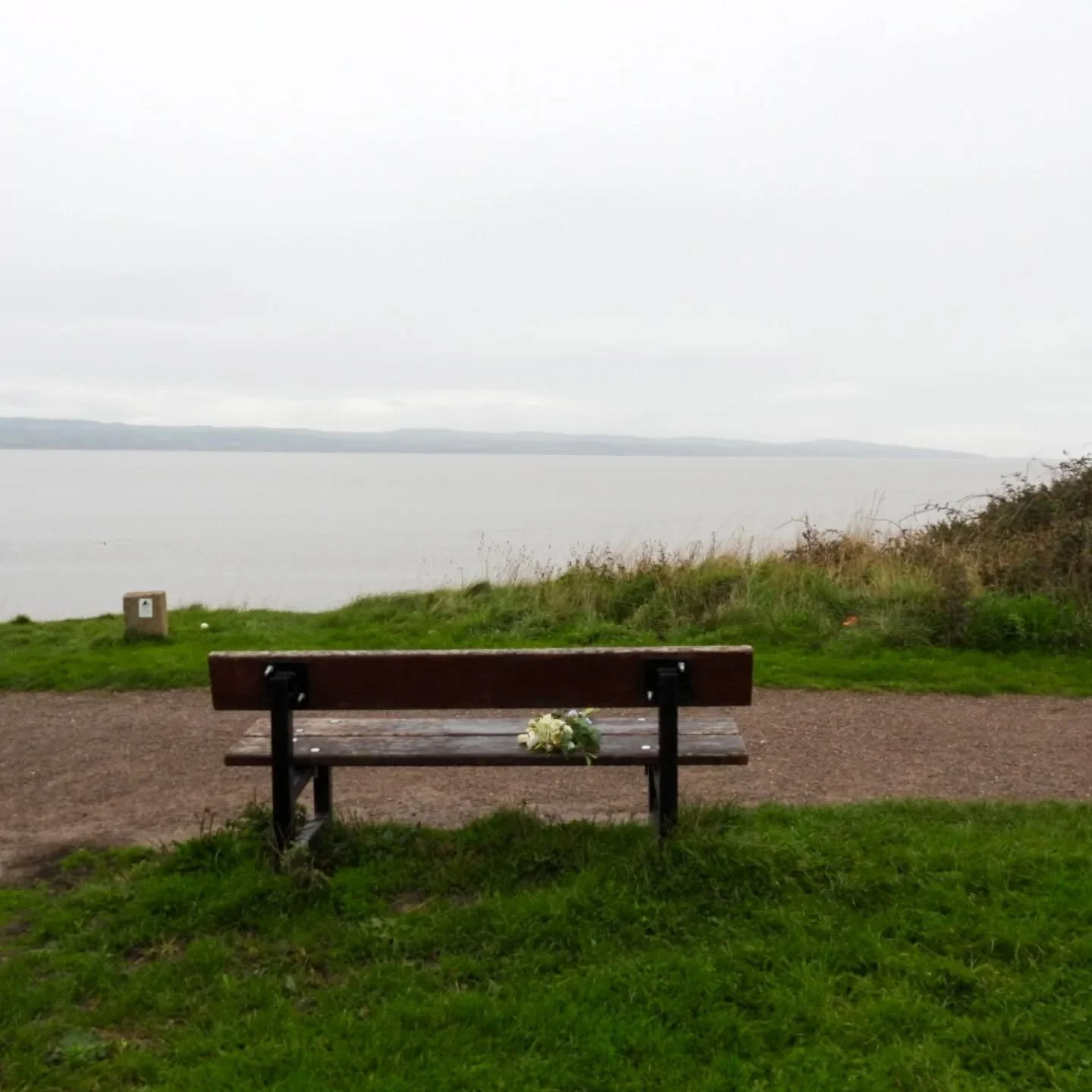 A wooden park bench with flowers resting on it, facing a large body of water under a cloudy sky.
