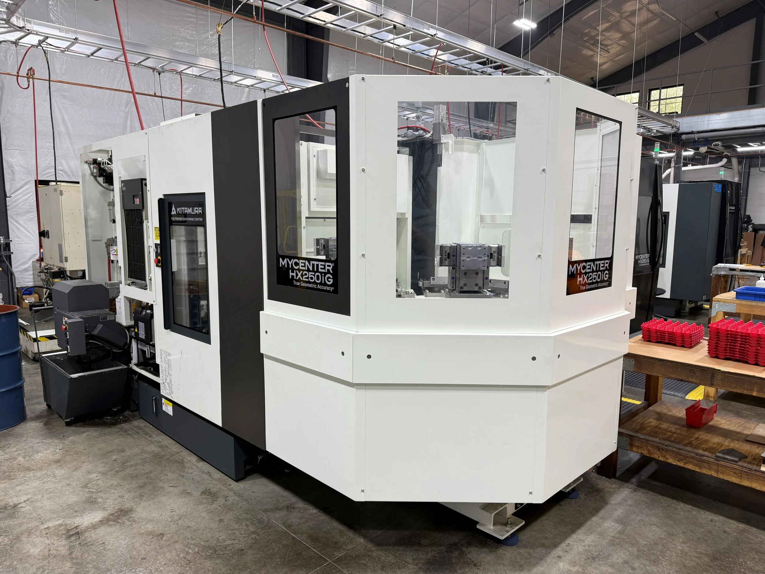 A large white industrial machine with black windows and labels reading 'MYCENTER HX250IG'. The machine is in a factory or workshop setting with tools and equipment around.