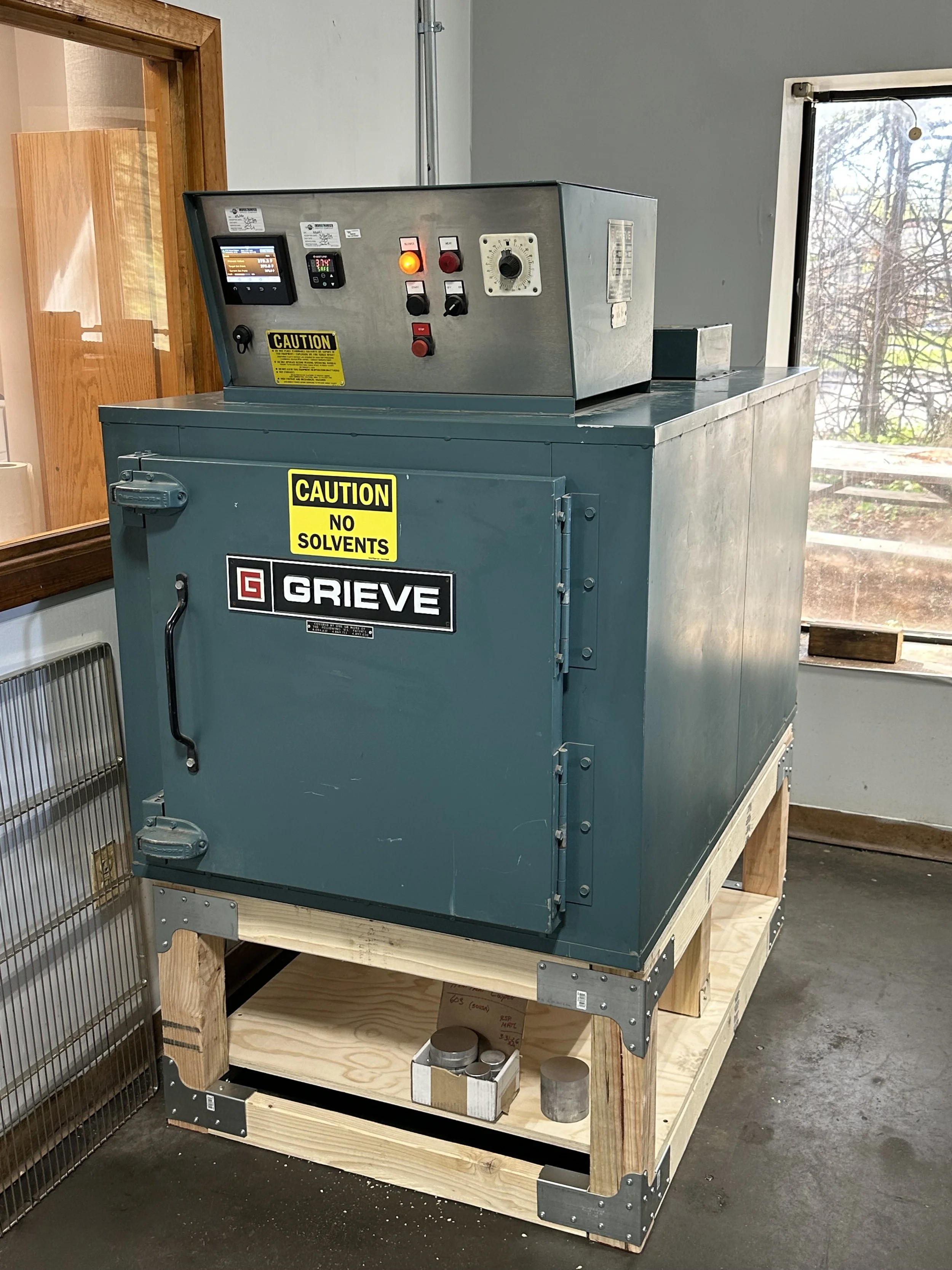 Metal industrial machine labeled 'Grieve' on a wooden pallet stand in a workshop. The machine has various control panels and warning stickers, with a window showing a tree outside.