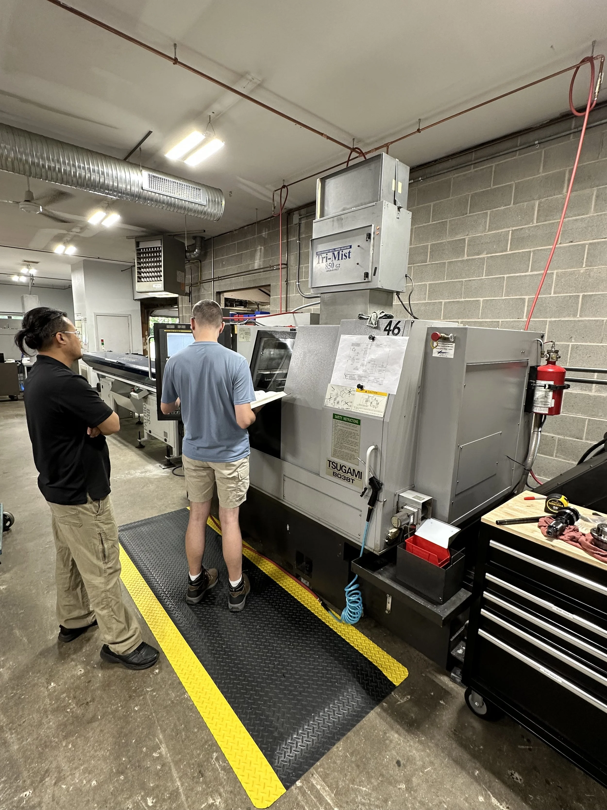 Two men are standing in front of a large industrial machine in a workshop environment, observing closely.