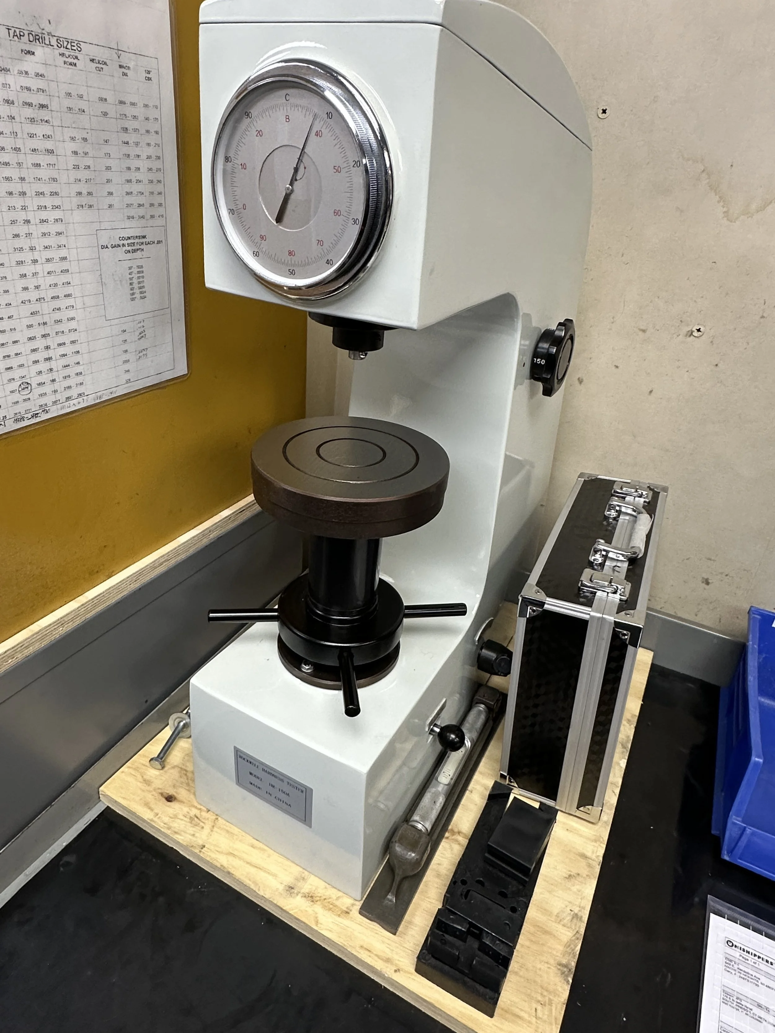 A precision vise mounted on a workbench with a balance scale and a black metal step block nearby. There is a chart on the wall and a metal box with unlabeled items to the right.