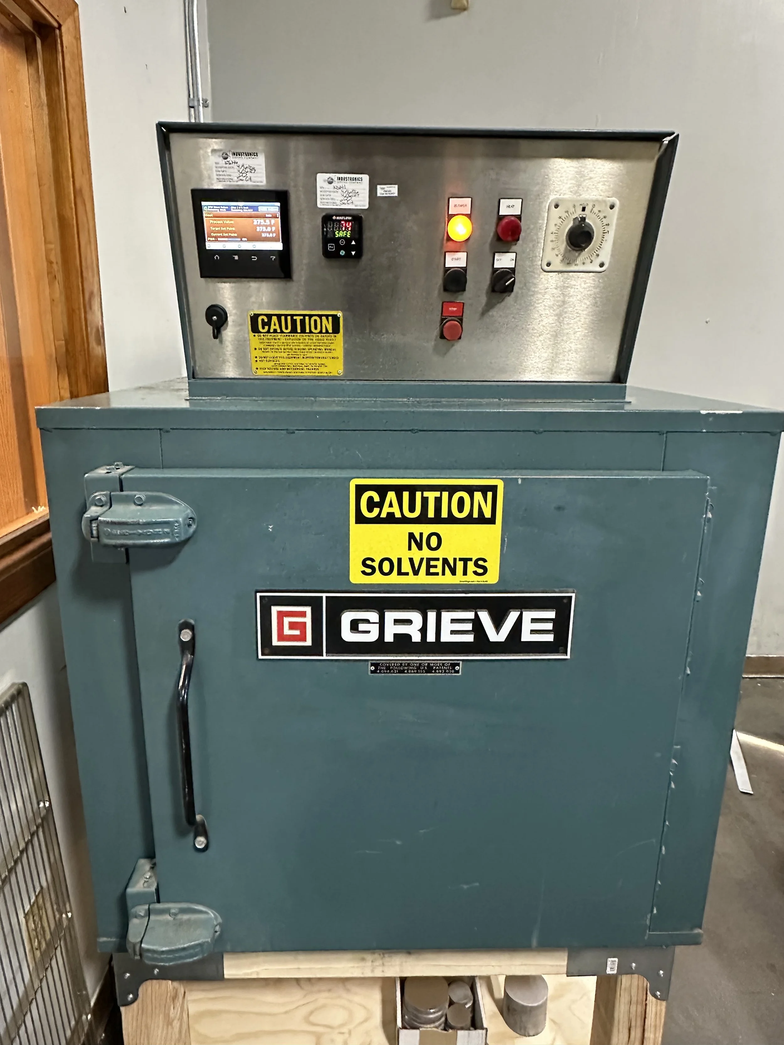 A metal industrial machine with warning labels and control knobs, sitting on a wooden pallet. The machine has a screen displaying information and a large gauge on the right side. The machine has a sign that says 'CAUTION NO SOLVENTS' and a brand label 'GRIEVE'.