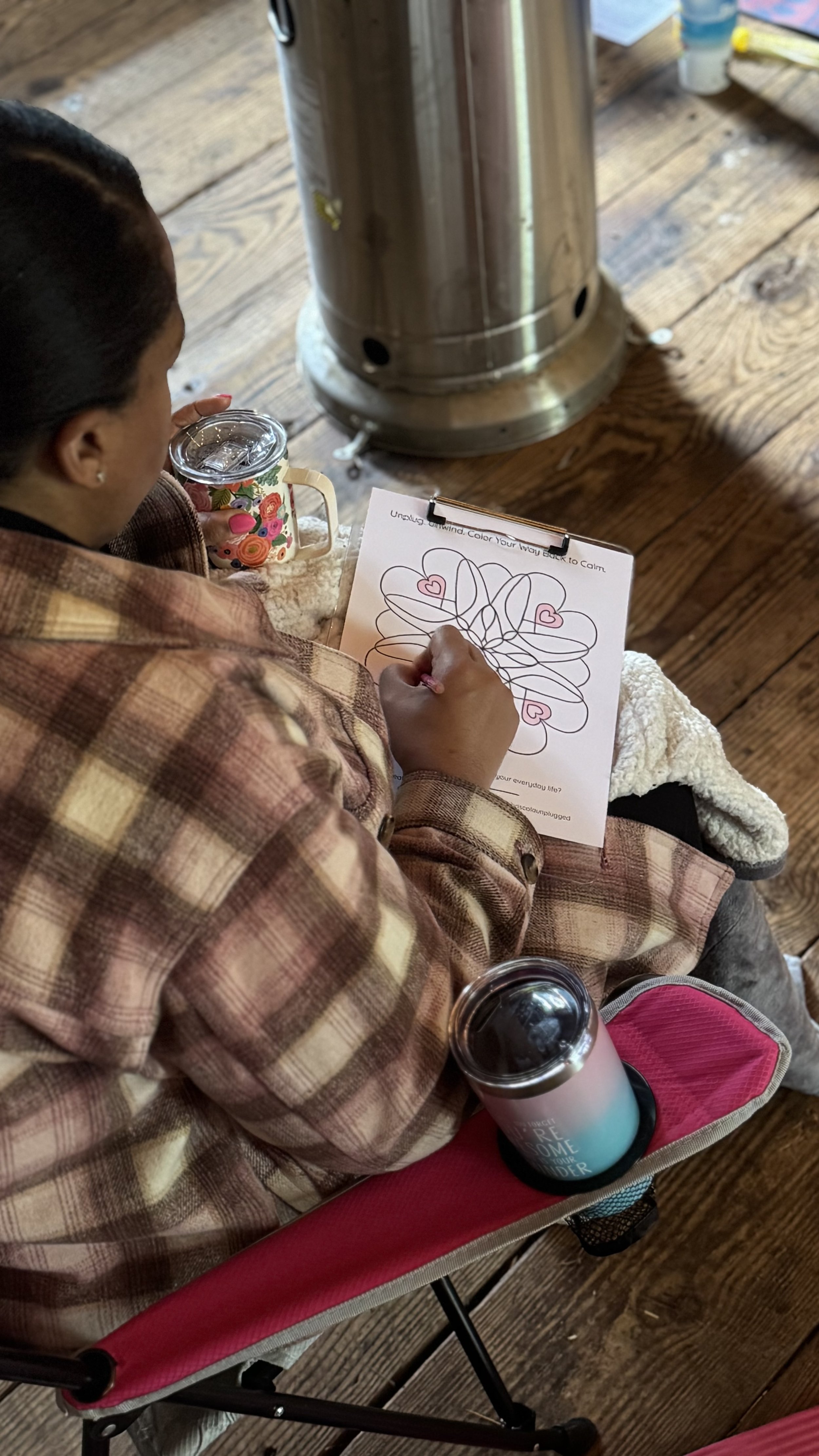 A woman sitting in a pink camping chair, coloring a heart-themed pattern on paper with a black marker. She has a water bottle in the cup holder of the chair, and there is a large metal heater and some supplies on a wooden floor nearby.
