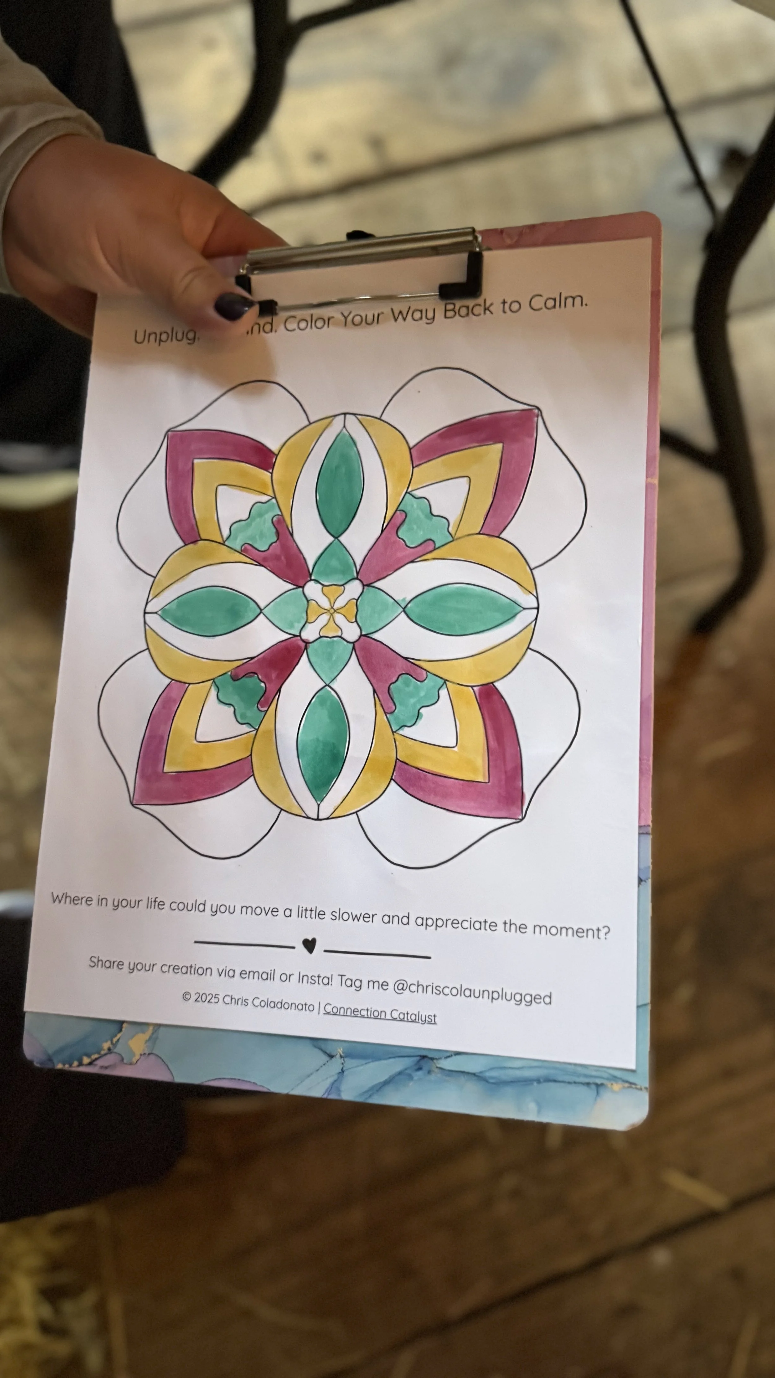 A person holding a coloring sheet of a symmetrical mandala design, partially colored with pink, green, and yellow. The sheet has instructions about unplugging and coloring to return to calm, and a prompt encouraging reflection on moving slower and appreciating the moment. The bottom also contains social media tags and a copyright notice.