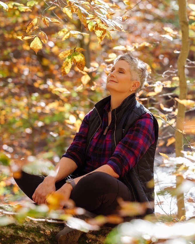 A woman sitting outdoors in a forest during autumn, smiling with her eyes closed, surrounded by yellow and orange leaves.