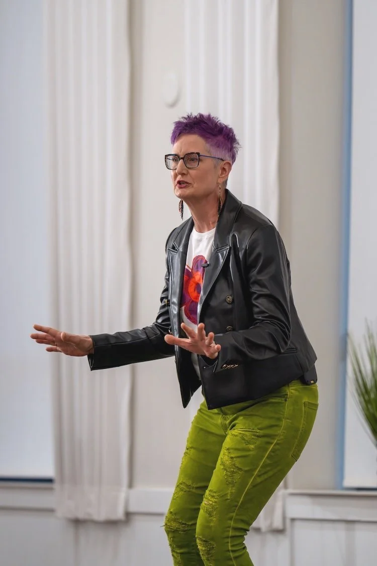 Person with purple hair, wearing glasses, a black leather jacket, graphic t-shirt, and green pants, speaking or performing gesture.