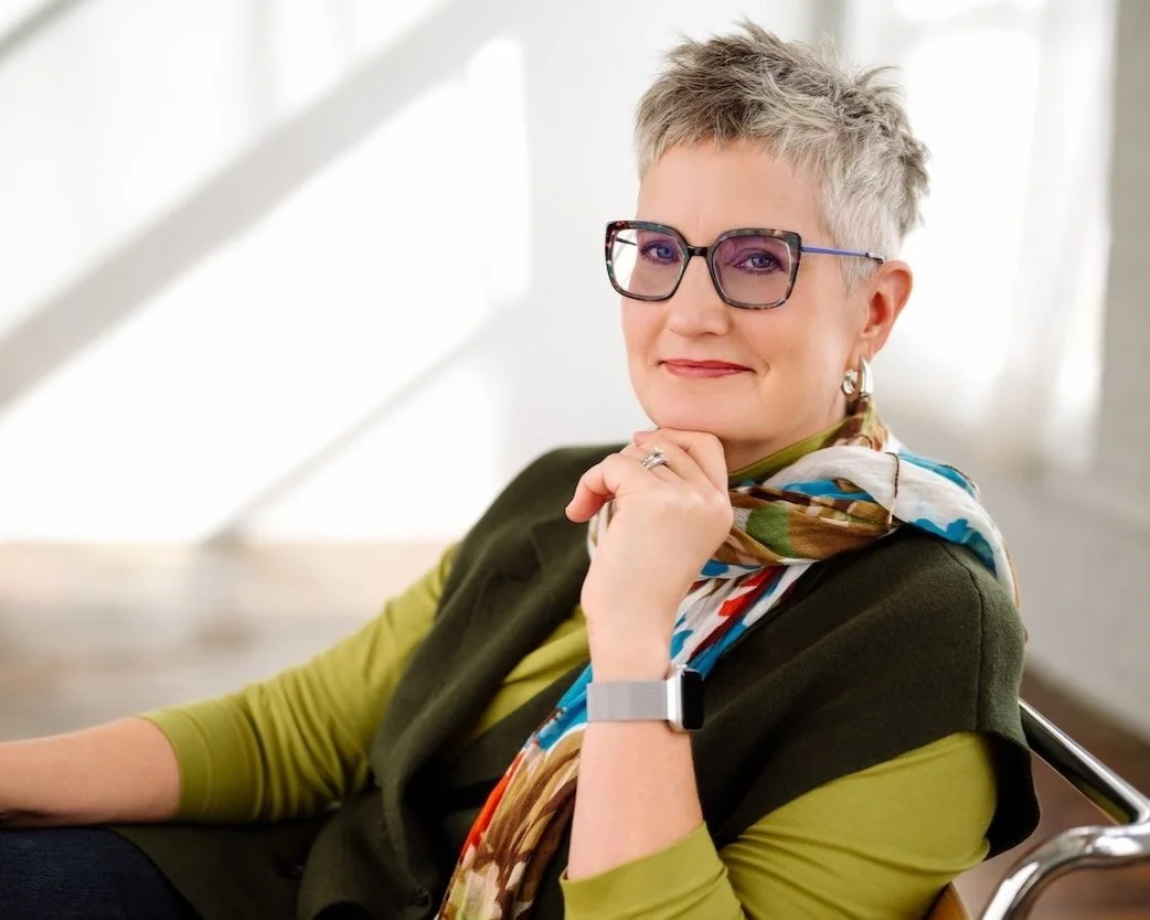 Woman with short hair, glasses, and a colorful scarf, wearing a green top and sitting indoors.