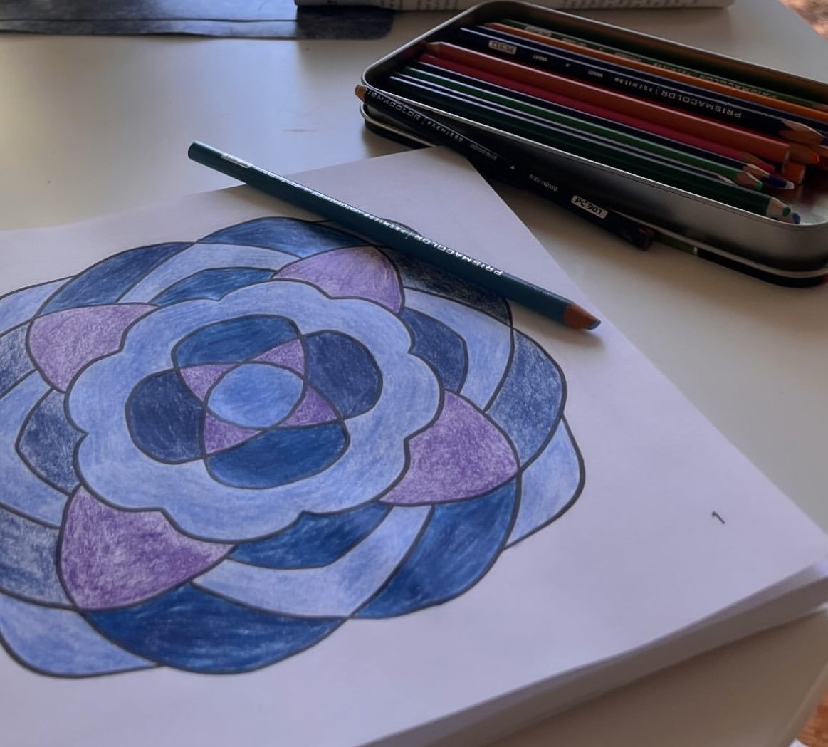 A coloring sheet with a geometric pattern in shades of blue and purple, colored with colored pencils, with a pencil resting on it. A box of colored pencils is open on the table.