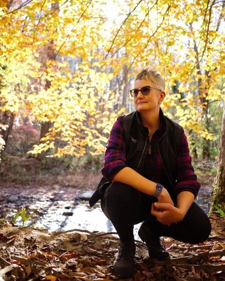 Person wearing glasses and casual clothing, crouching in a forest surrounded by autumn foliage.