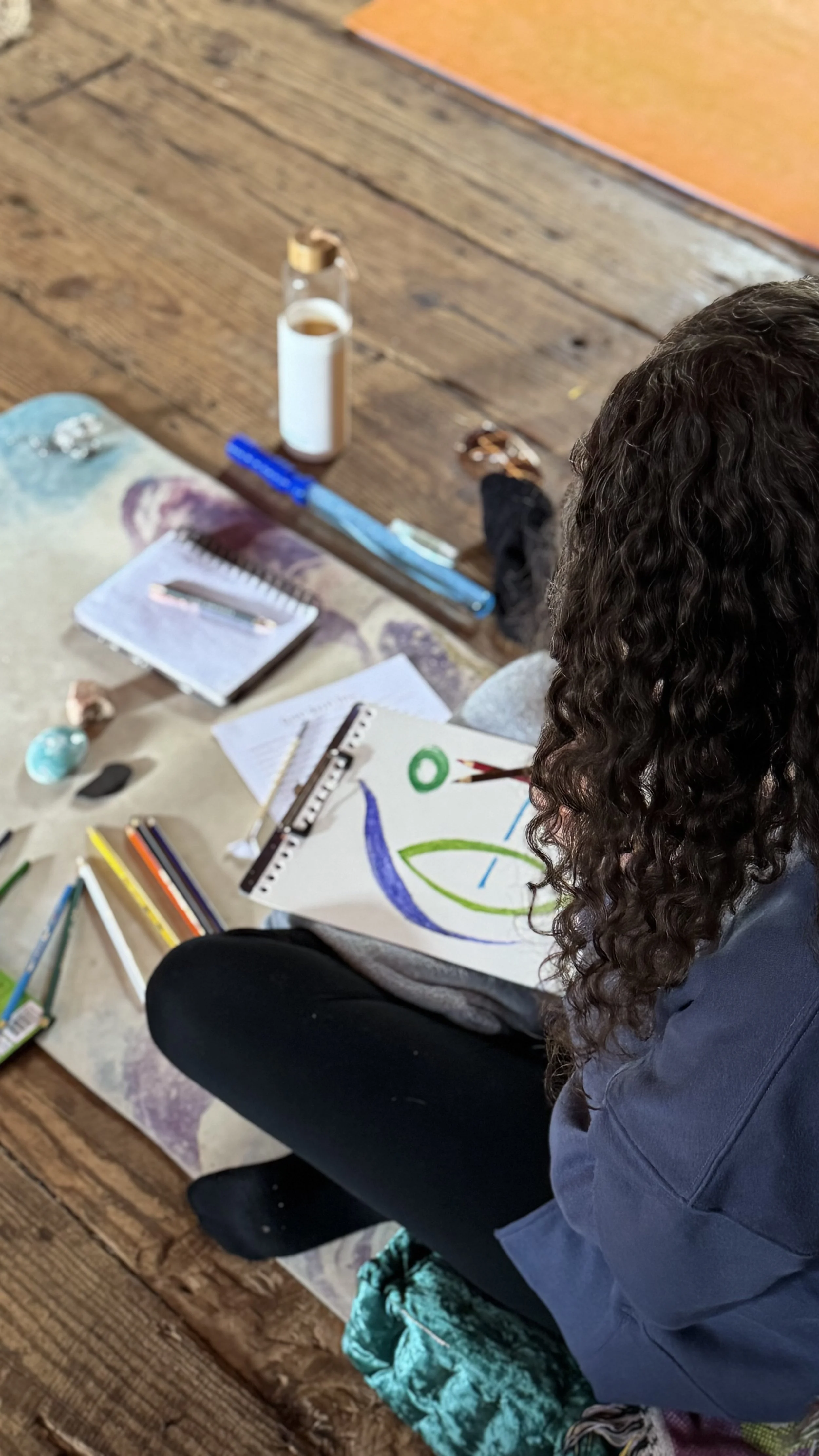 A person with curly hair drawing and coloring on a white sheet of paper, surrounded by various art supplies on a wooden floor.