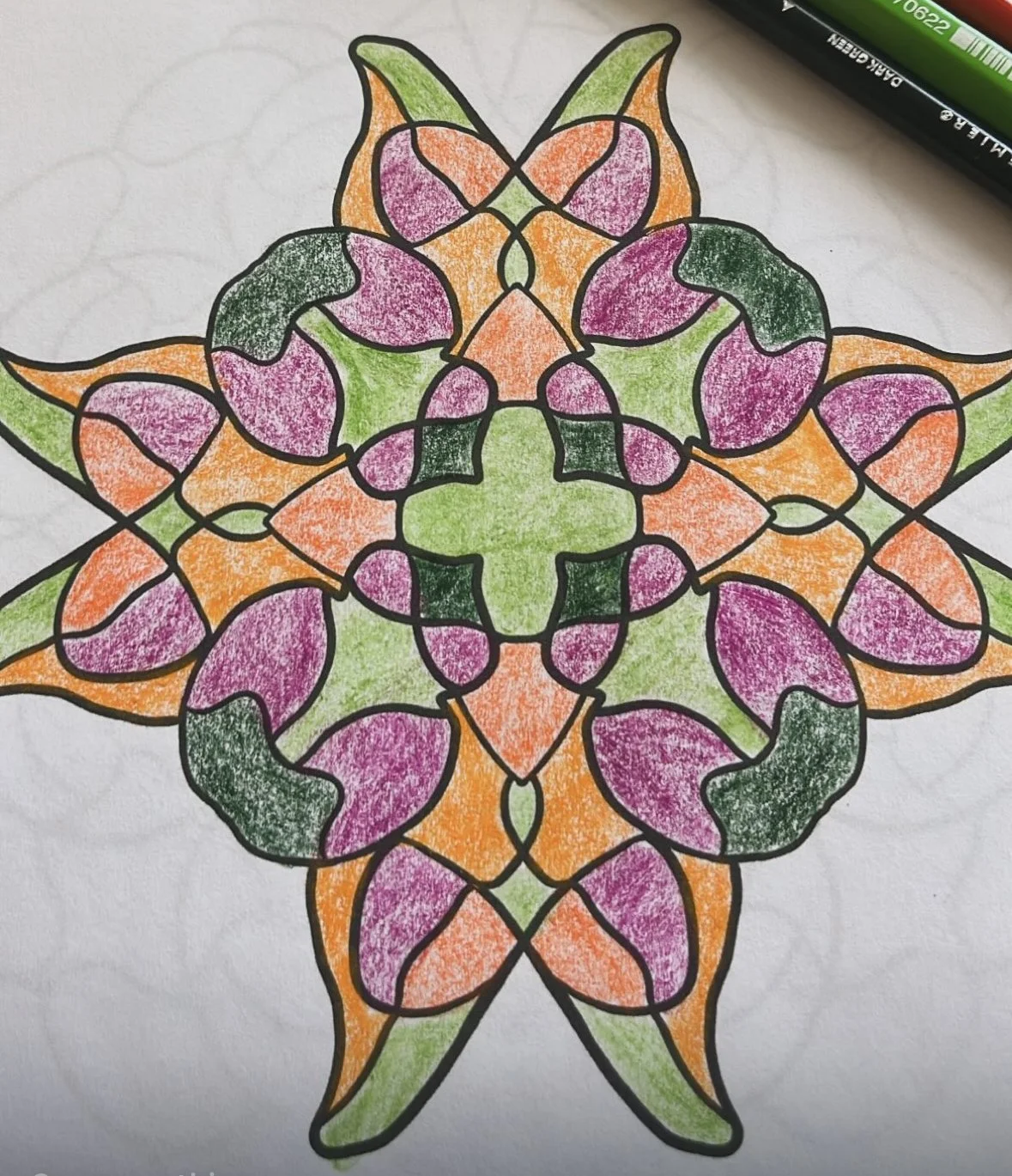 Colorful symmetrical mandala pattern drawn with black outlines, filled with shades of green, pink, orange, and purple, on white paper, with a black and green marker nearby.