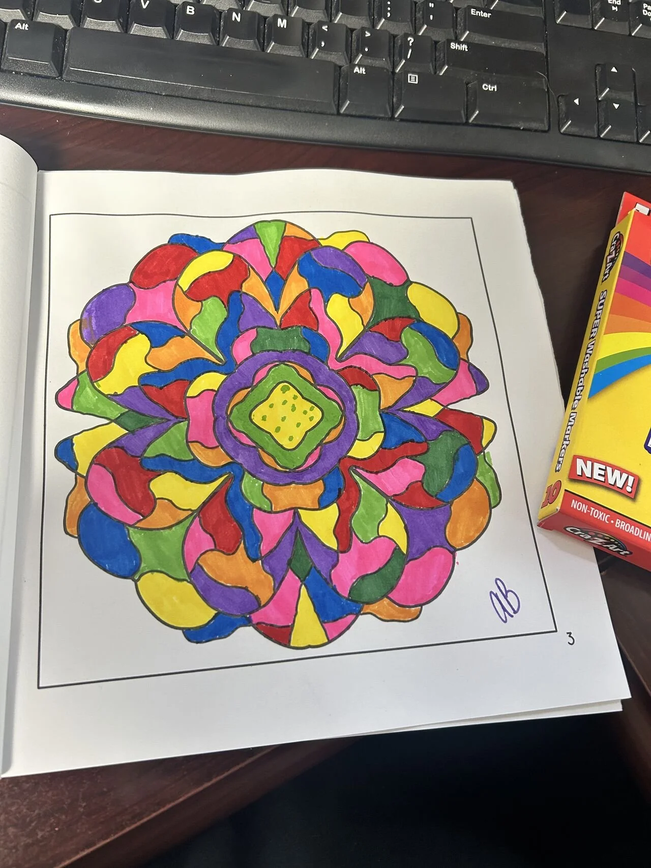 Colorful mandala coloring page on paper, with a box of crayons nearby, on a desk with a keyboard in the background.