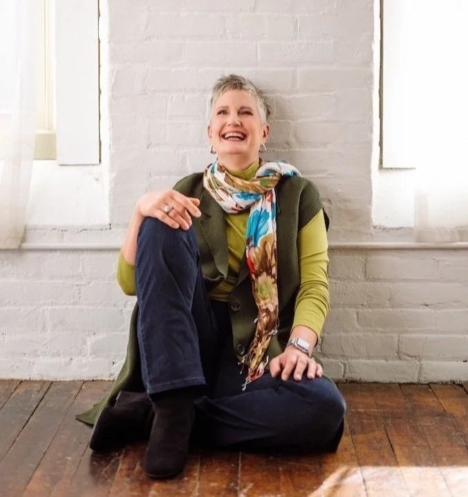 Person sitting on the floor in front of a white brick wall, wearing a green sweater, floral scarf, and dark jeans, smiling and laughing.