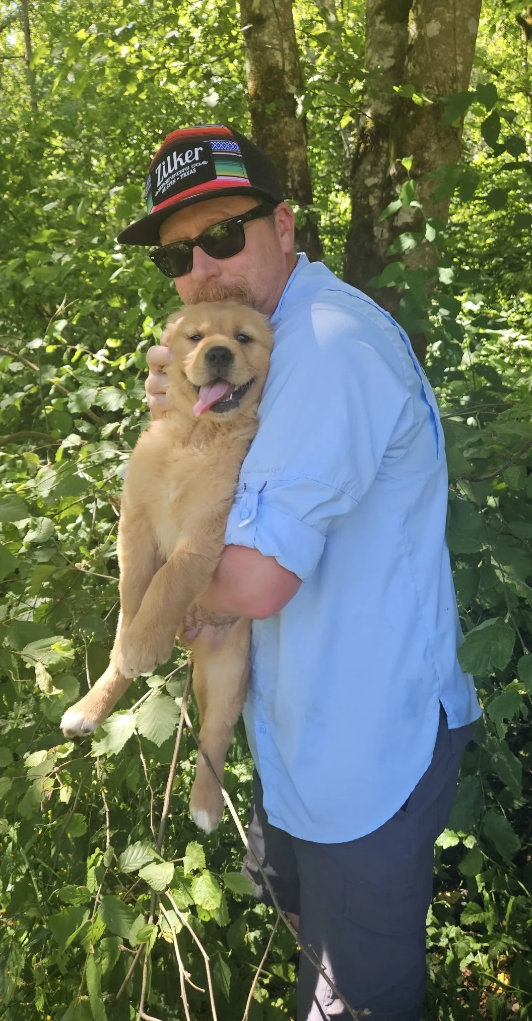 Man wearing sunglasses and a hat holding a fluffy puppy outdoors surrounded by greenery.