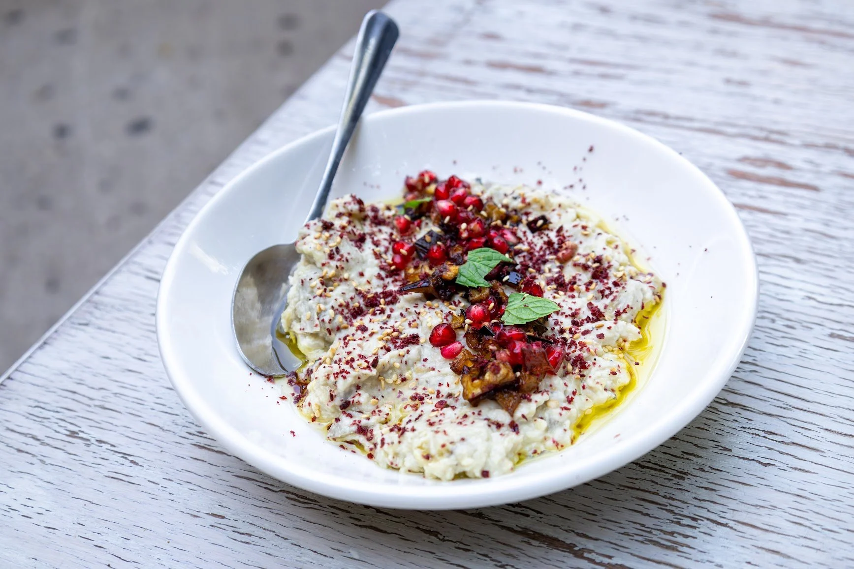 A white bowl containing a dish topped with pomegranate seeds, herbs, and spices, with a spoon resting on the side, placed on a weathered white wooden table.