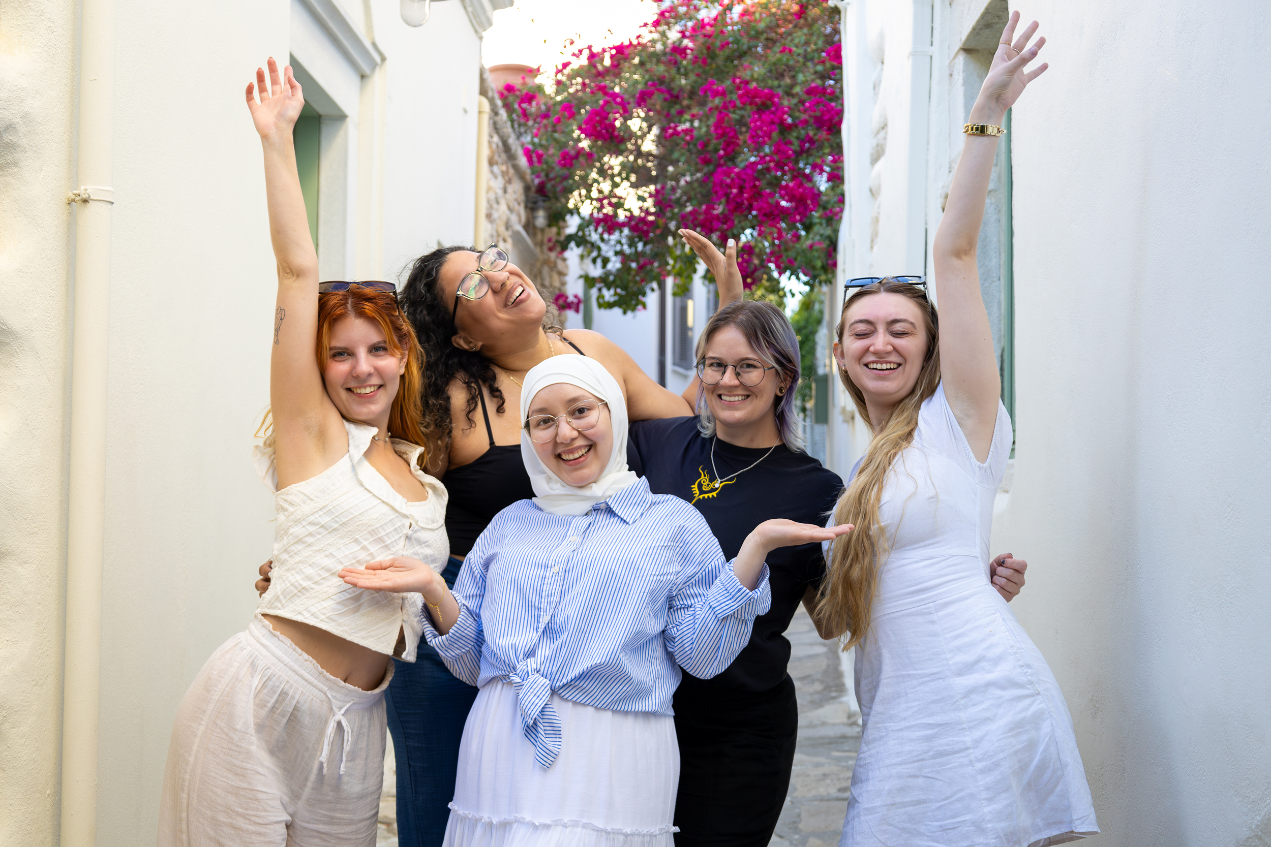 Group of five women of diverse backgrounds smiling and celebrating together outdoors in a narrow street with flowering plants in the background.