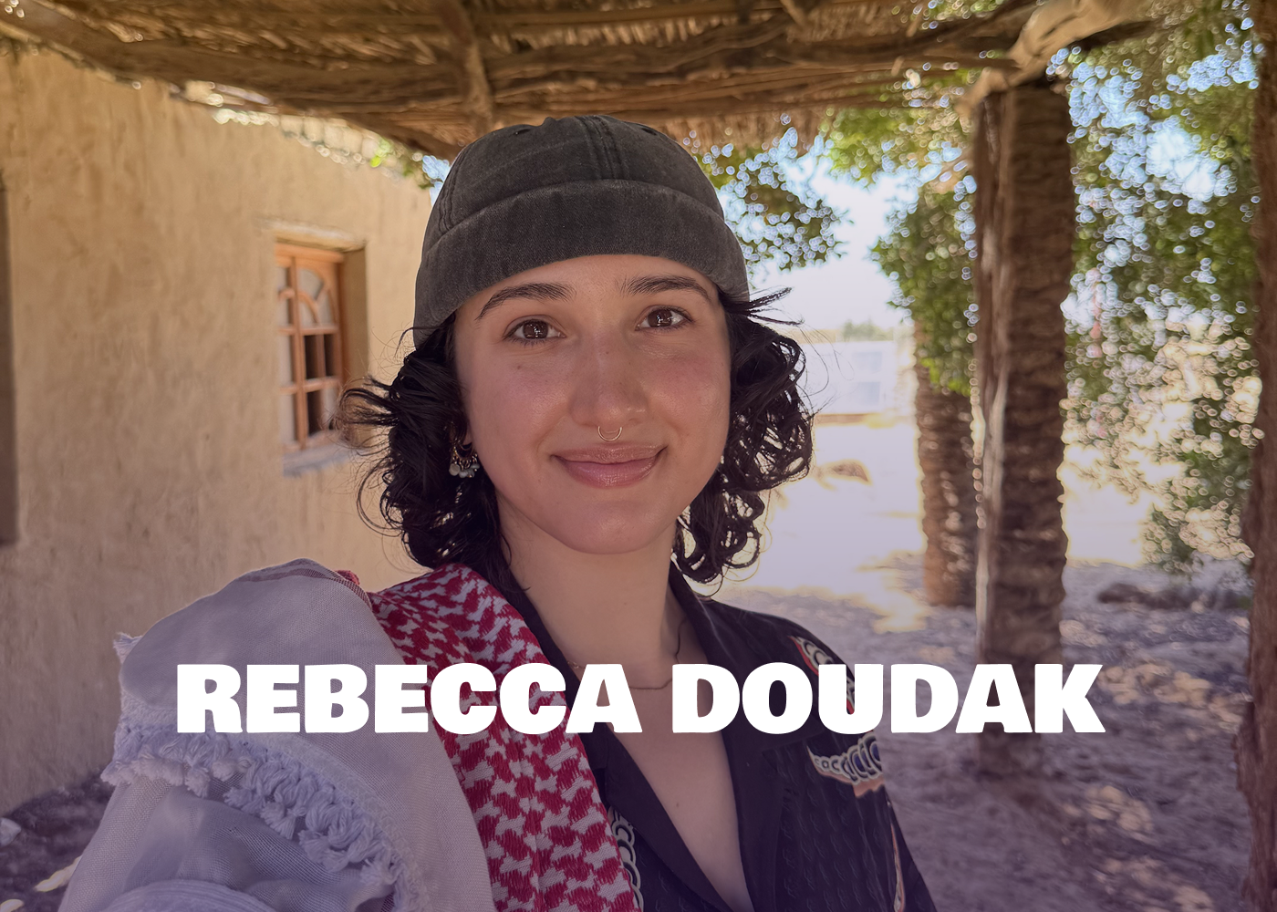 Portrait of a woman with dark curly hair, wearing a gray beanie, earring, and a patterned scarf, standing outside under a wooden pergola with trees and a building in the background.
