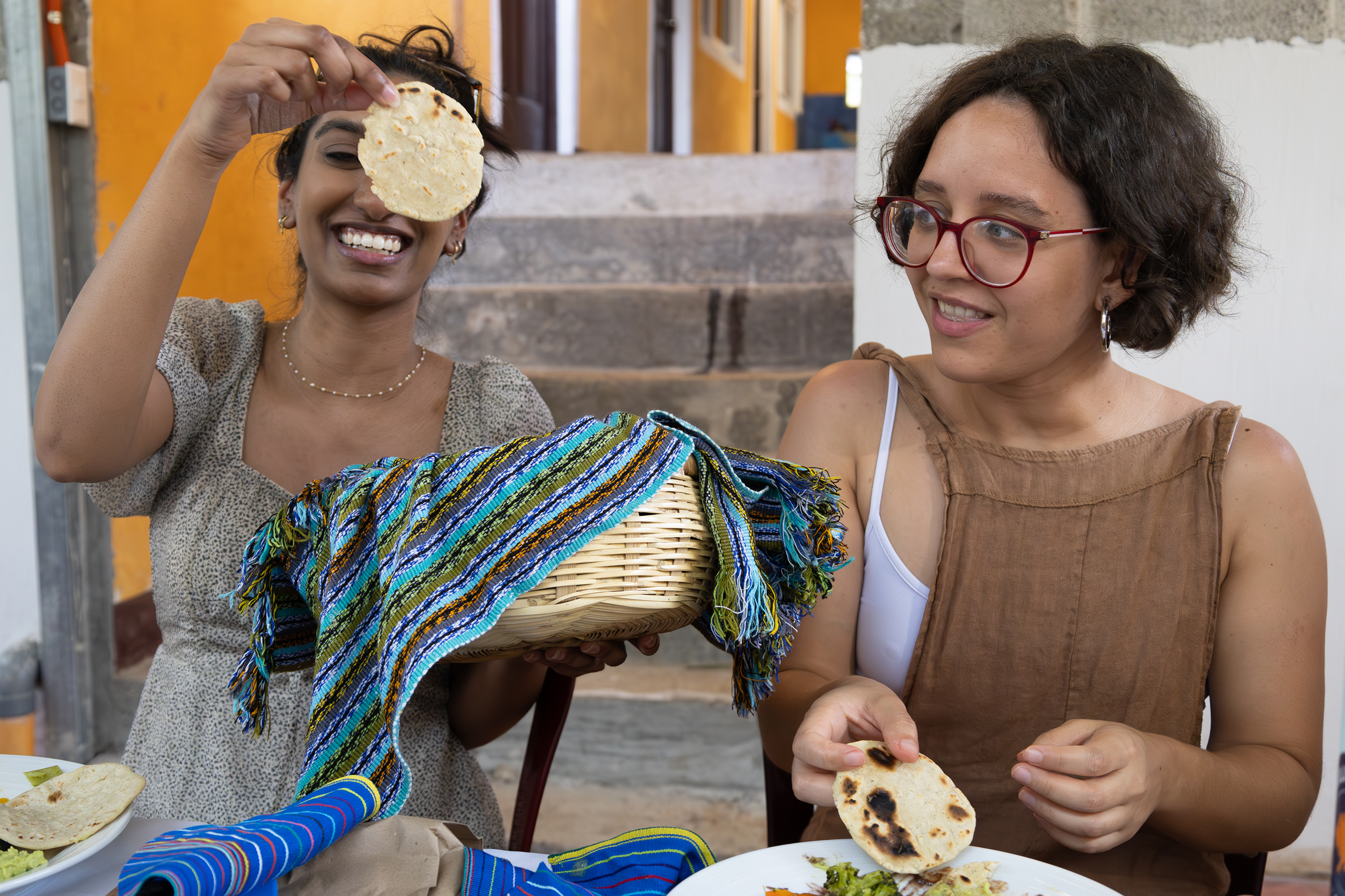 Two women enjoying Mexican food with tortillas and toppings, with one woman holding a tortilla in front of her face and smiling, while the other woman looks on smiling.