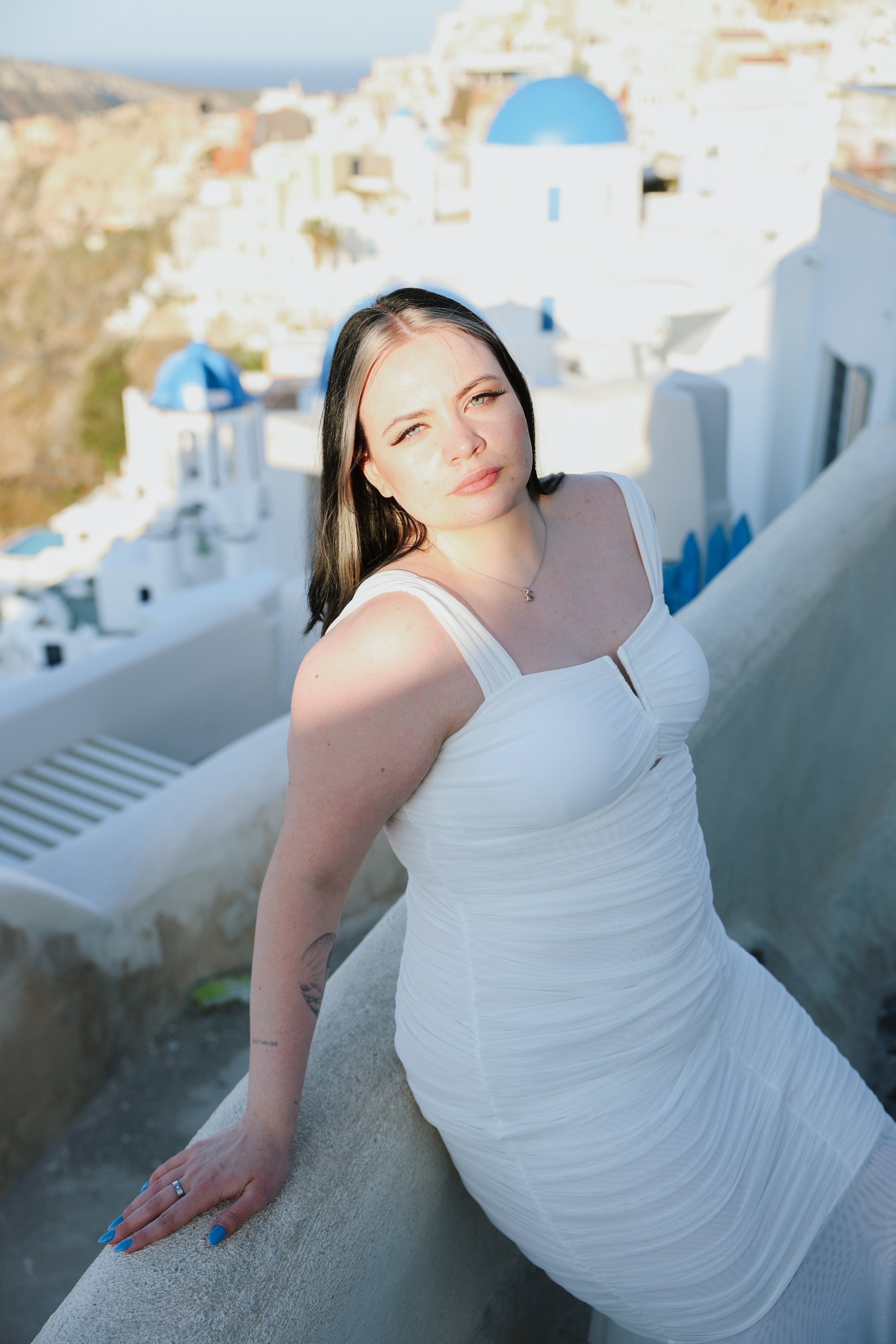 A woman with long dark hair and fair skin, wearing a white dress, sitting on a ledge against a background of classic white buildings with blue domes, resembling Santorini, Greece.