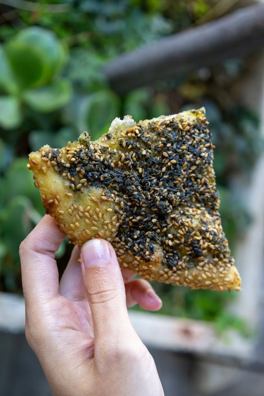 Close-up of a hand holding a piece of bread with sesame and black sesame seeds on top, with a blurred green outdoor background.