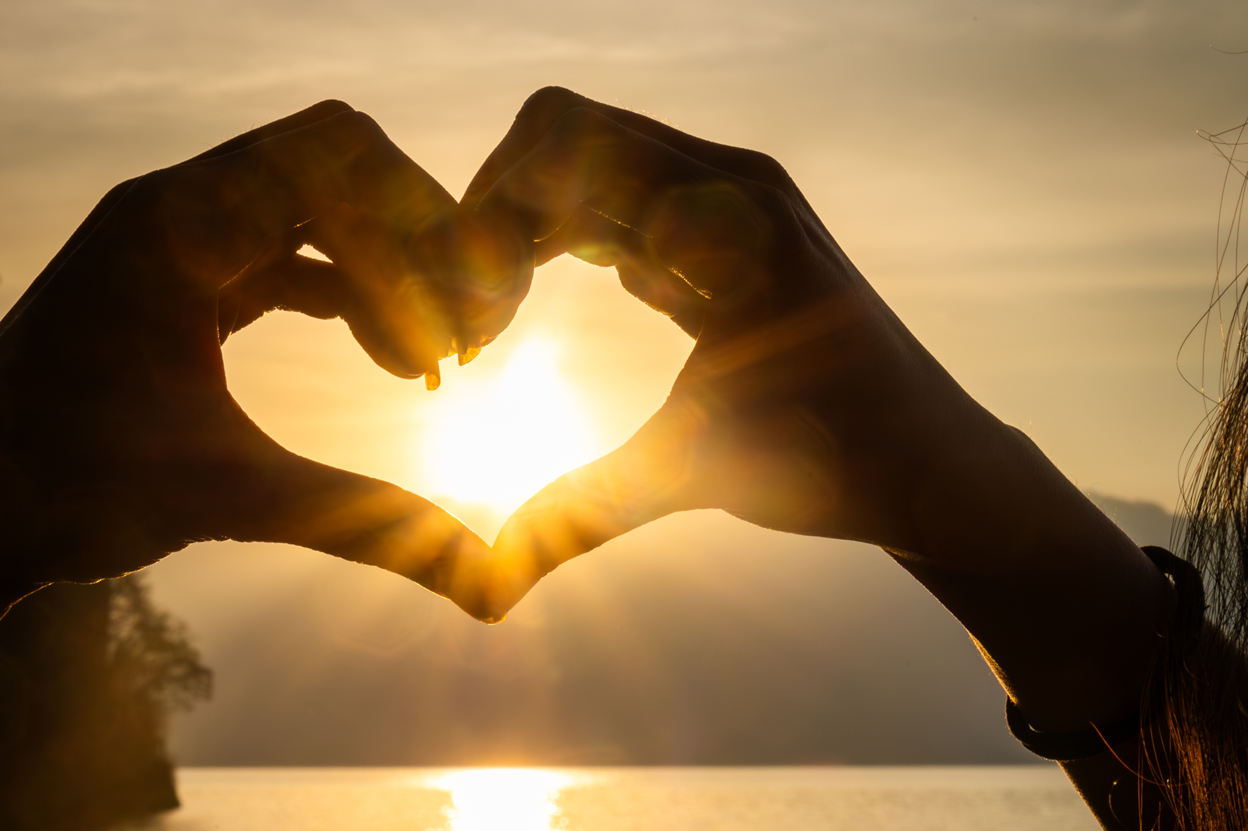 Two hands forming a heart shape around the setting sun by a body of water at sunset.
