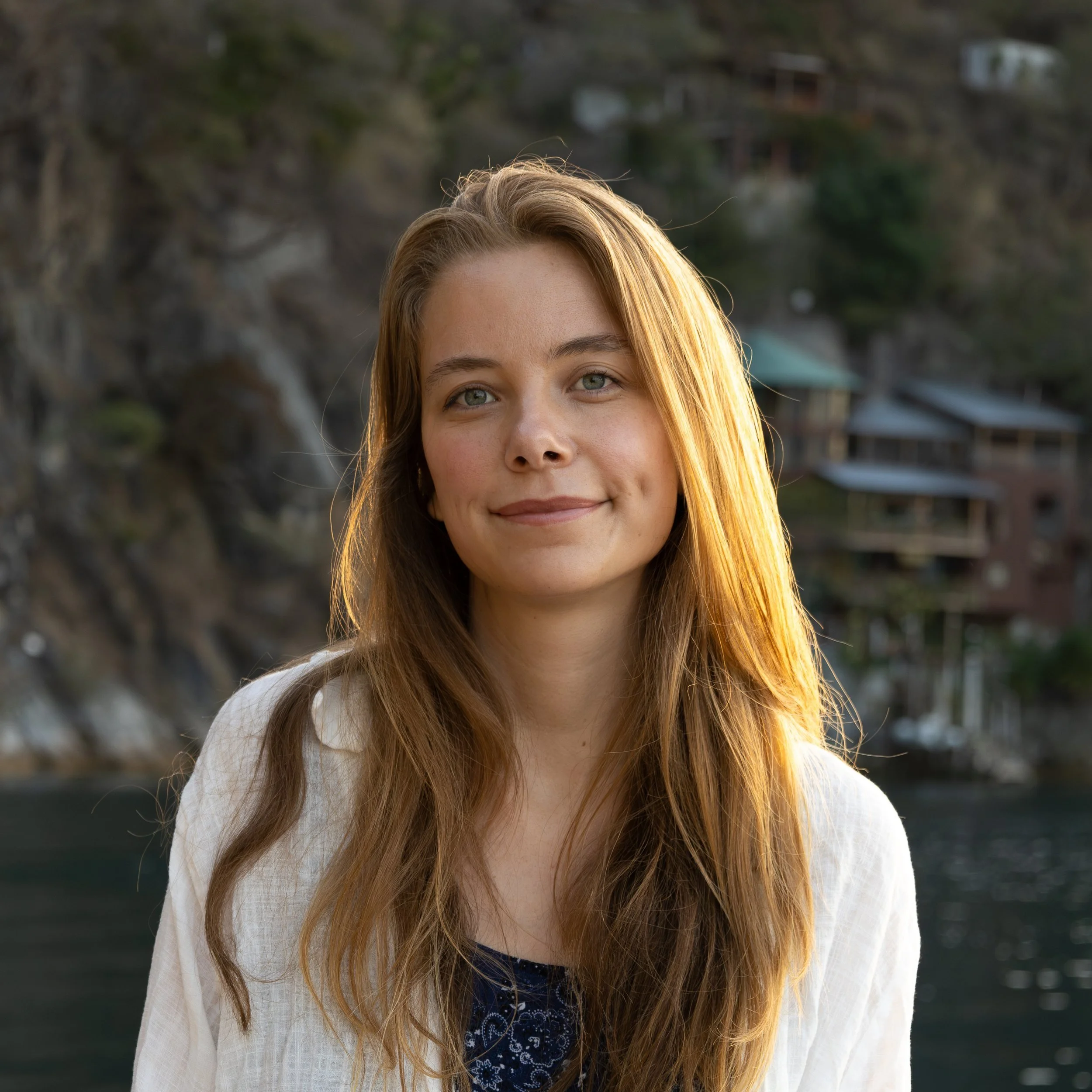 A young woman with long, wavy red hair and blue eyes, smiling softly, outdoors with a rocky hillside and houses in the background.