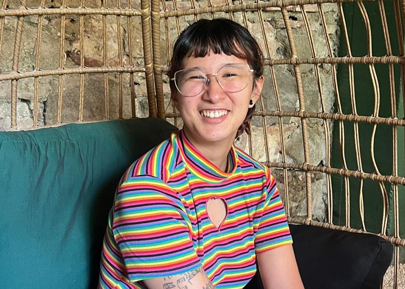 A smiling person with short dark hair, glasses, and a rainbow striped shirt, sitting on teal and green seating with a bamboo-style lattice and stone wall background.