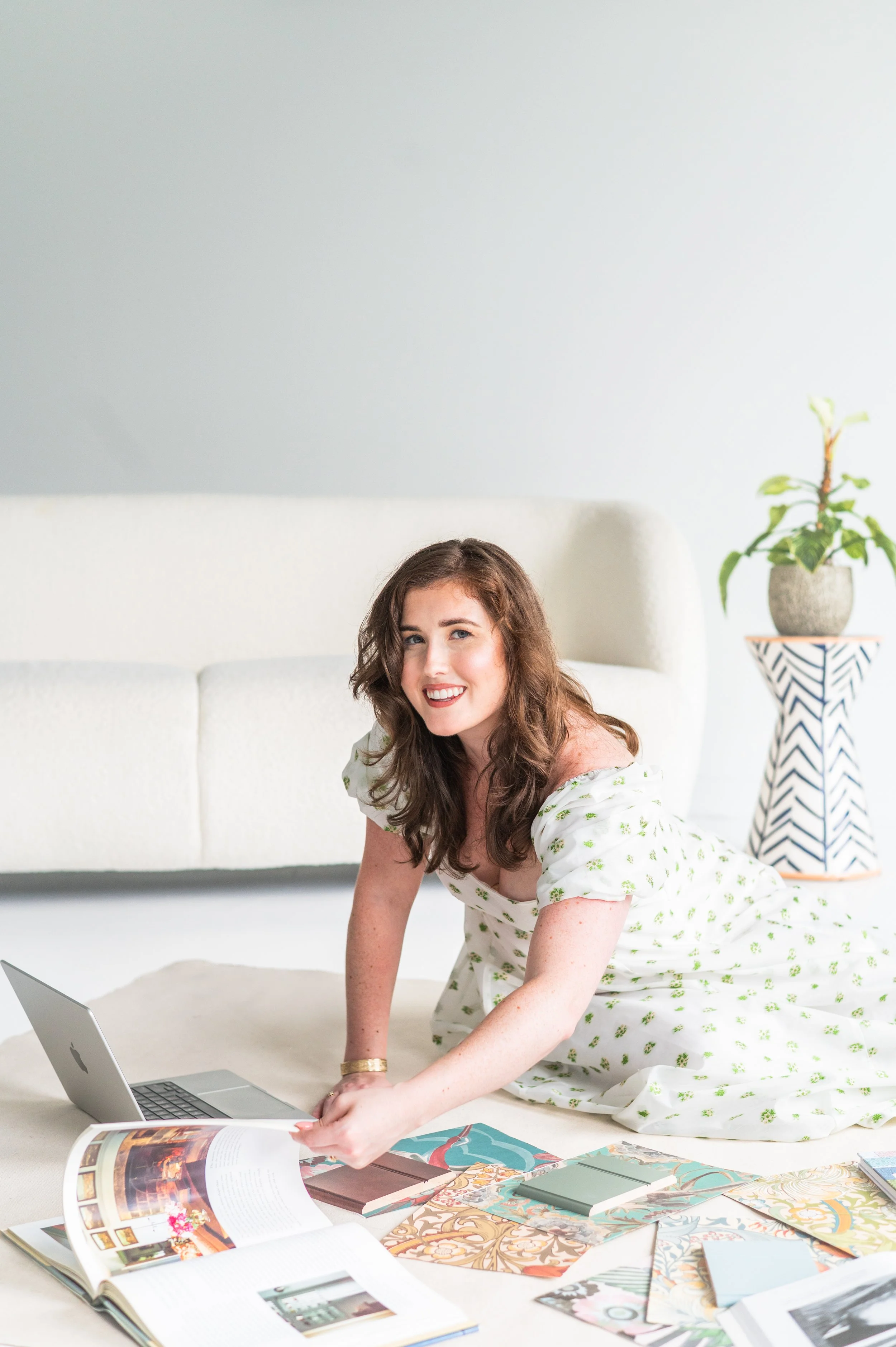 A woman with brown hair, wearing a white dress with green floral patterns, is kneeling on the floor in front of a table covered with colorful books and magazines. She is smiling at the camera, with a laptop open in front of her and a potted plant on a stand in the background.