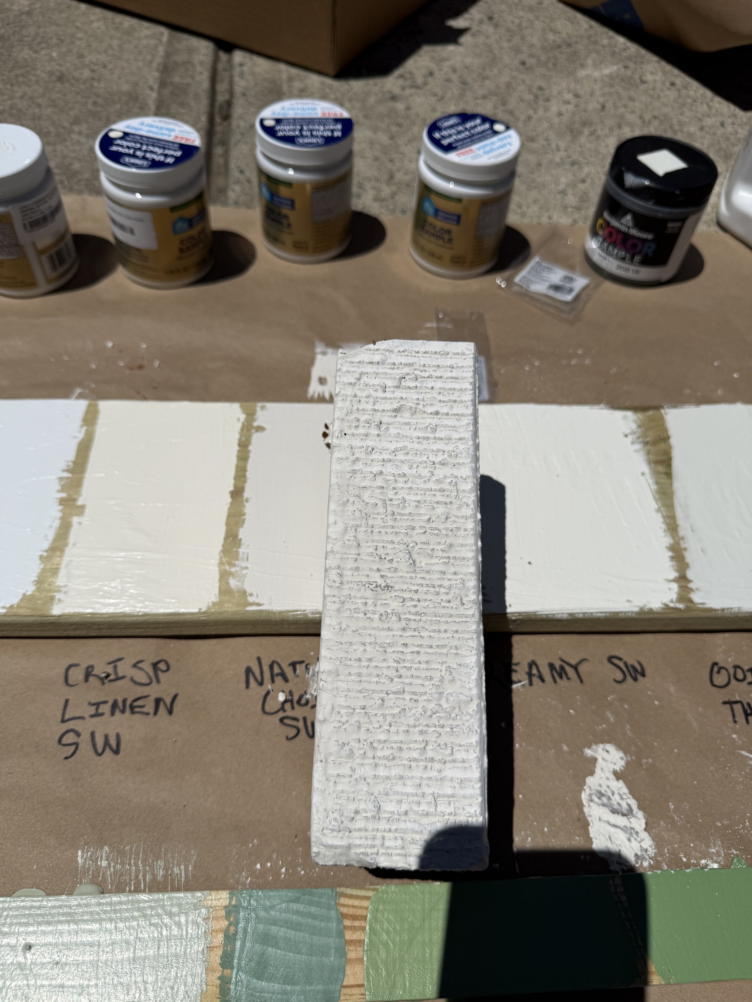 Several shades of white and green paint are visible on two pieces of scrap wood.  In this scene, a limewashed spare brick is placed on top of the wood with white paint swatches.