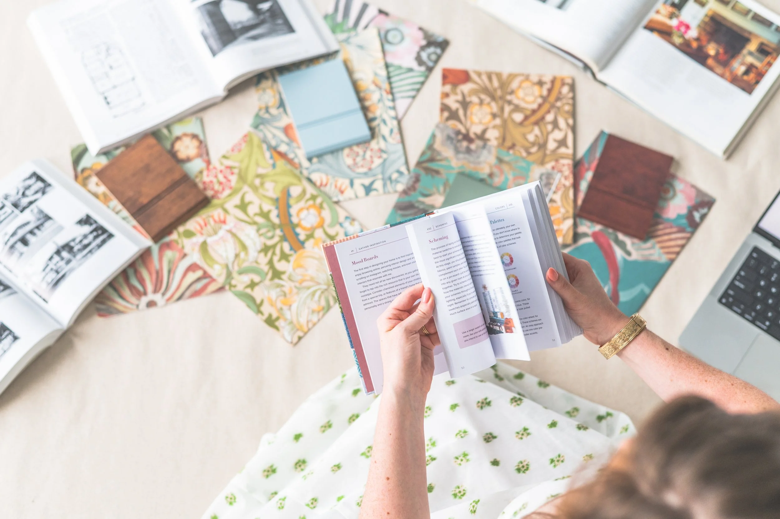 Person holding an open booklet with design ideas, surrounded by color swatches, magazines, and a laptop on a light surface.