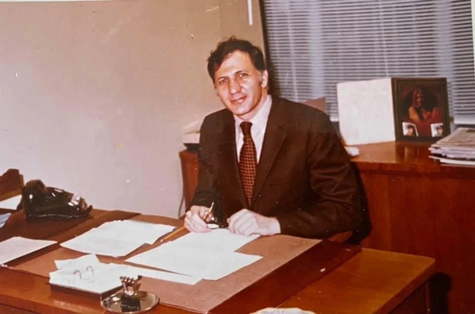 An Italian American gentleman sitting at a midcentury deck in an office building signing paperworks likely around the mid 70s