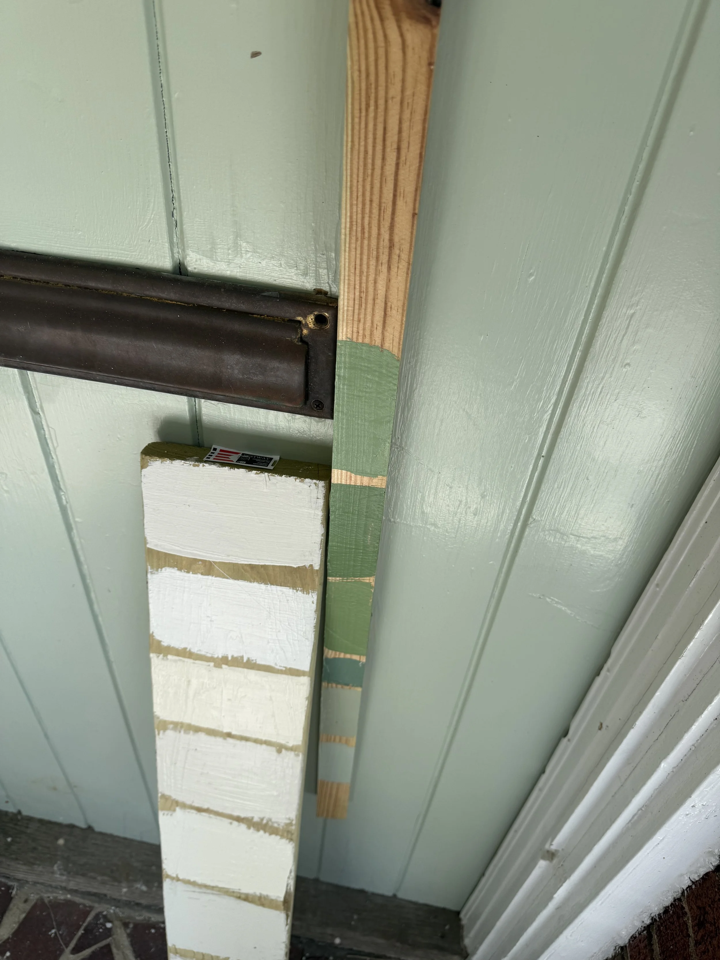 Paint swatches on scrap wood are seen leaning against a front door near a mail slot.