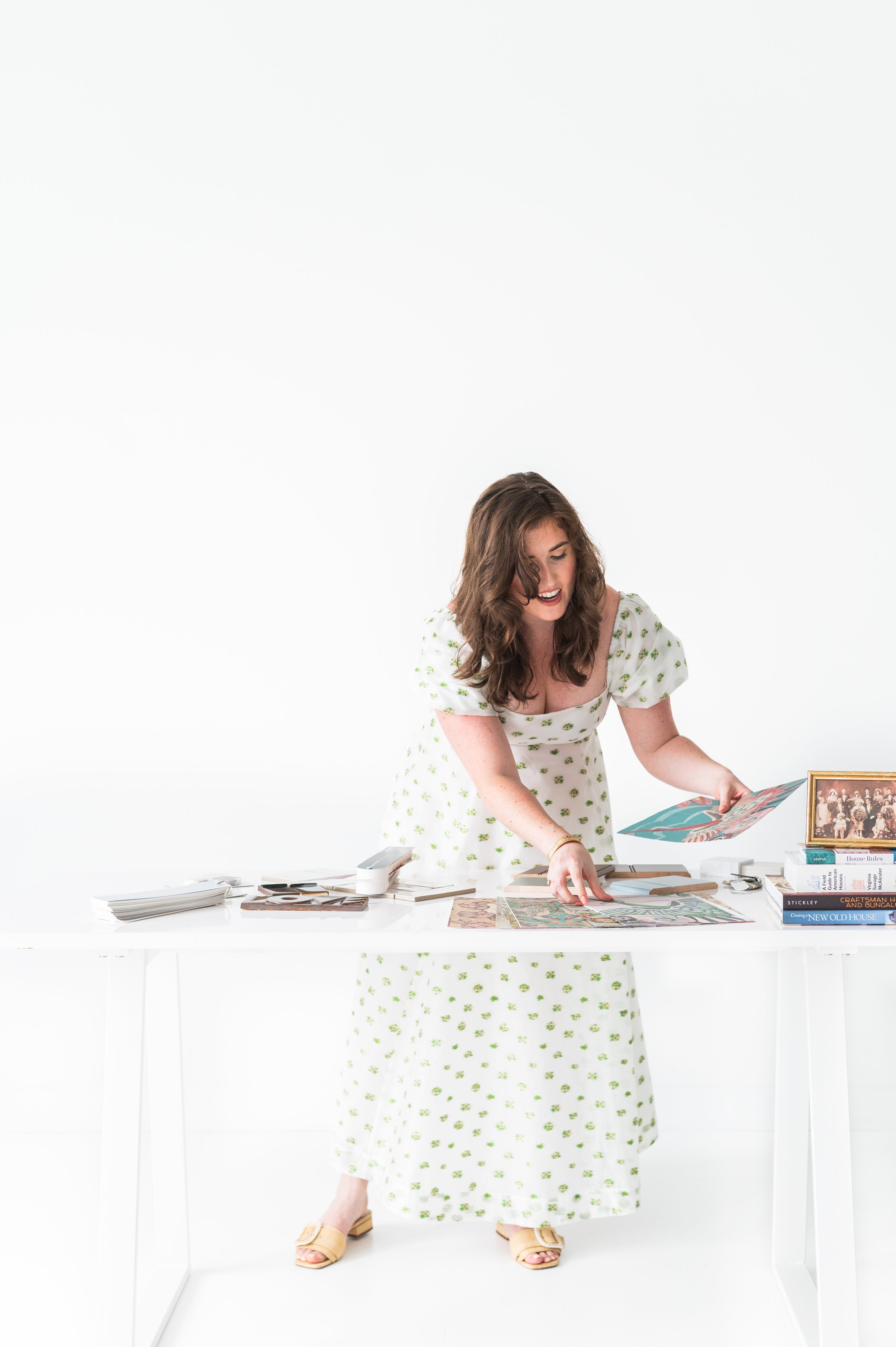 A woman with wavy brown hair in a white dress with green floral pattern is organizing colorful artwork on a white table in a bright white room. There are stacks of books, papers, and picture frames on the table.