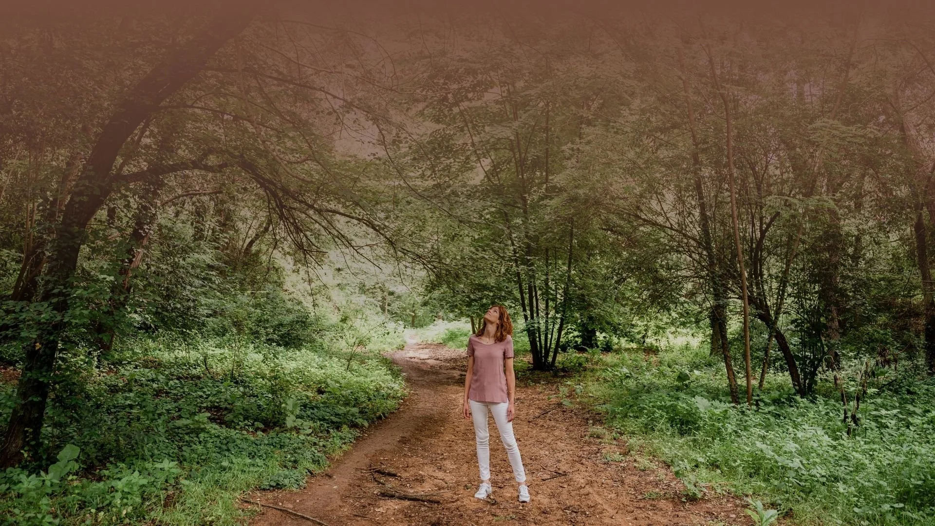 Mujer de pie en un sendero en un bosque con árboles verdes y follaje, mirando hacia arriba.
