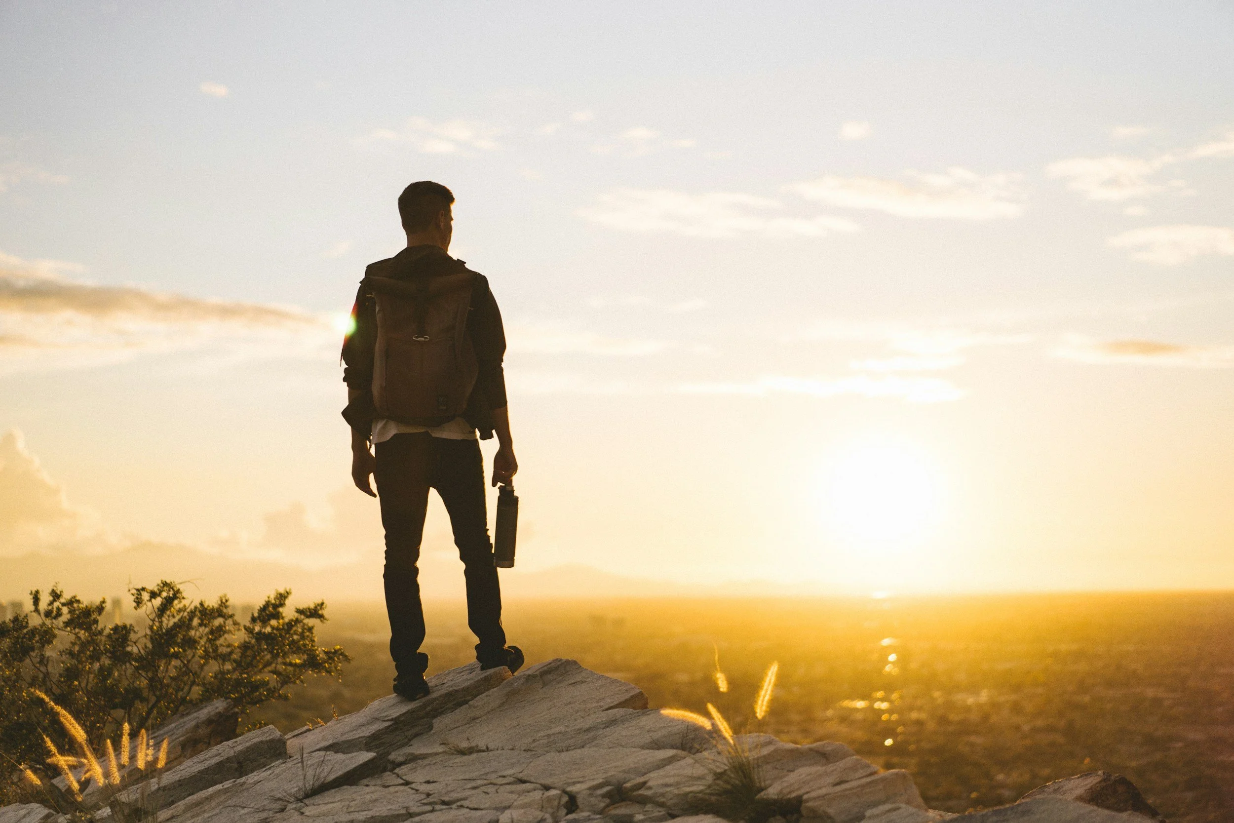 A man with a backpack standing on a rocky ledge at sunset, overlooking a vast landscape and ocean.