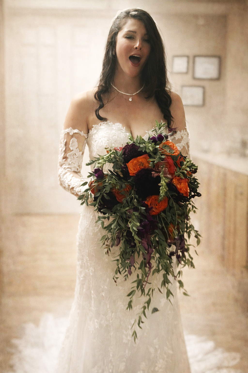A woman in a wedding dress holding a colorful bouquet of flowers, standing indoors with a neutral background.