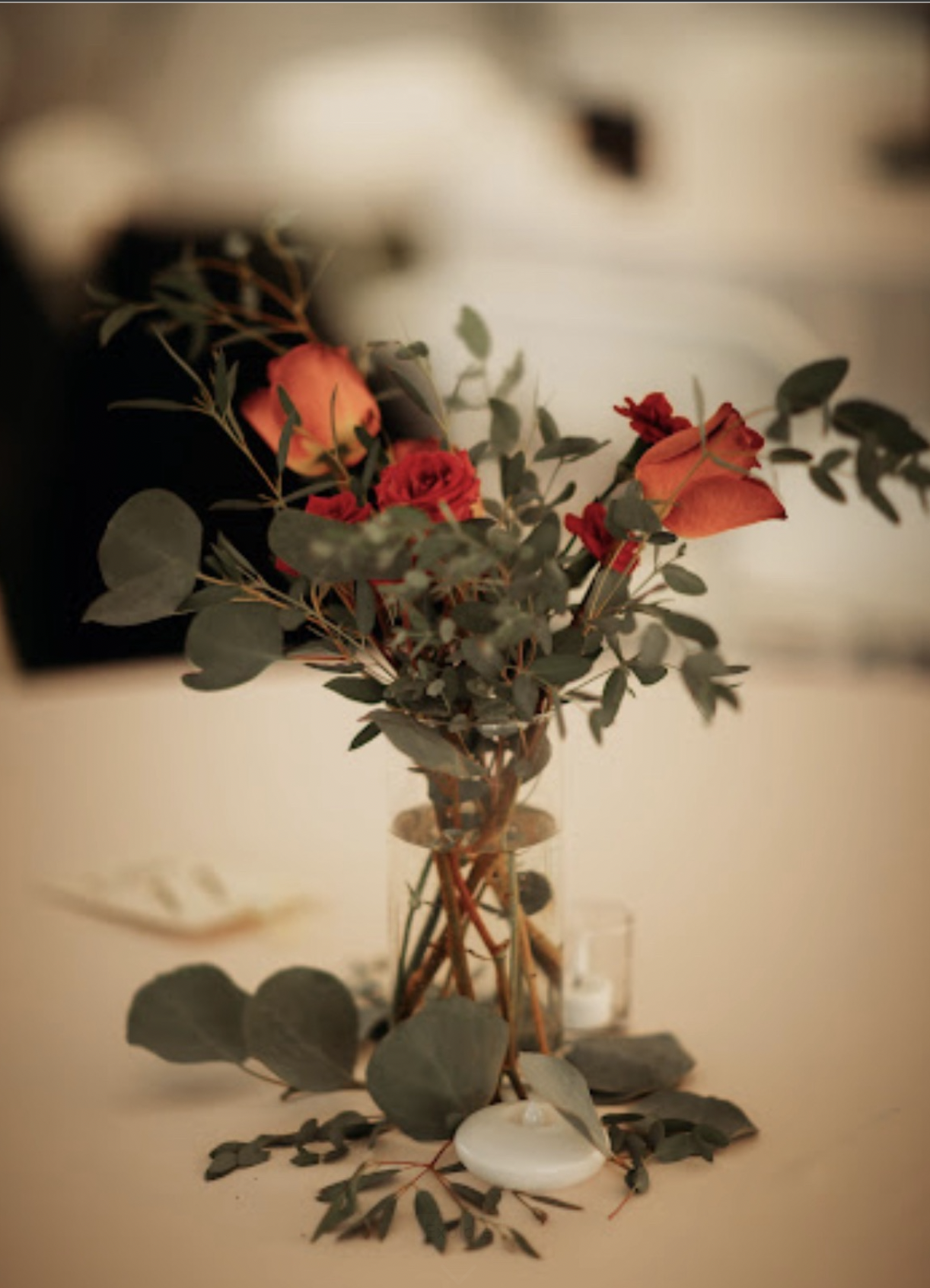 A bouquet of red and orange flowers with green leaves in a clear glass vase on a table, with scattered leaves and a white candle in front.