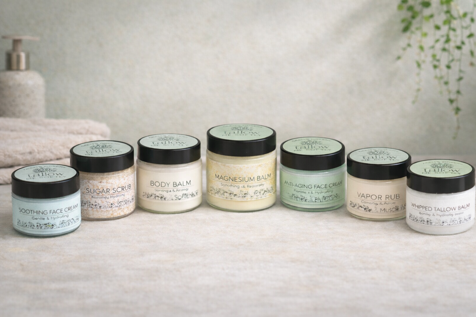 A collection of seven skincare balm jars with labels like Soothing Face Cream, Sugar Scrub, Body Balm, Magnesium Balm, Anti-Aging Face Cream, Vapor Rub, and Whipped Tallow Balm, arranged in a row on a beige surface with a textured background and a plant in the top right corner.