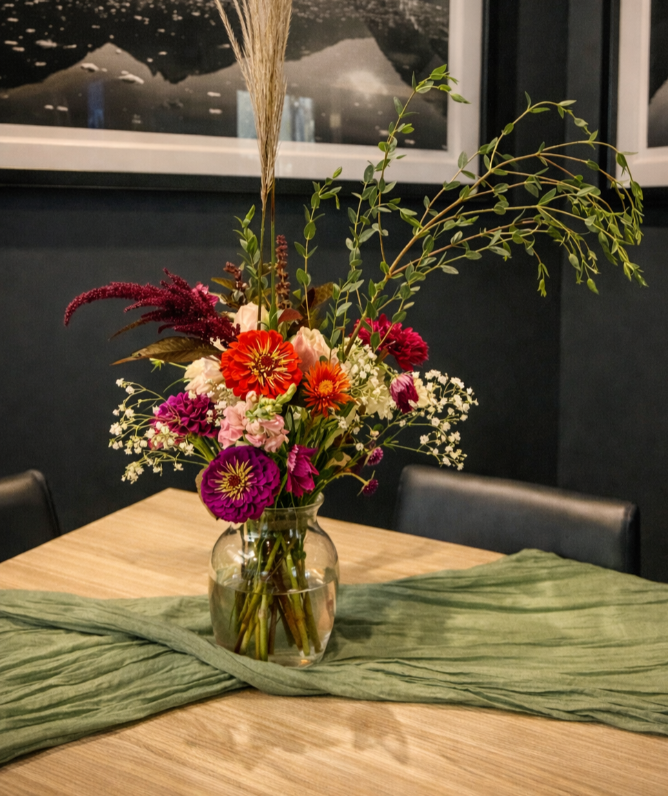A colorful floral arrangement in a glass vase on a wooden table with a green table runner, with framed nature photos on a dark wall in the background.